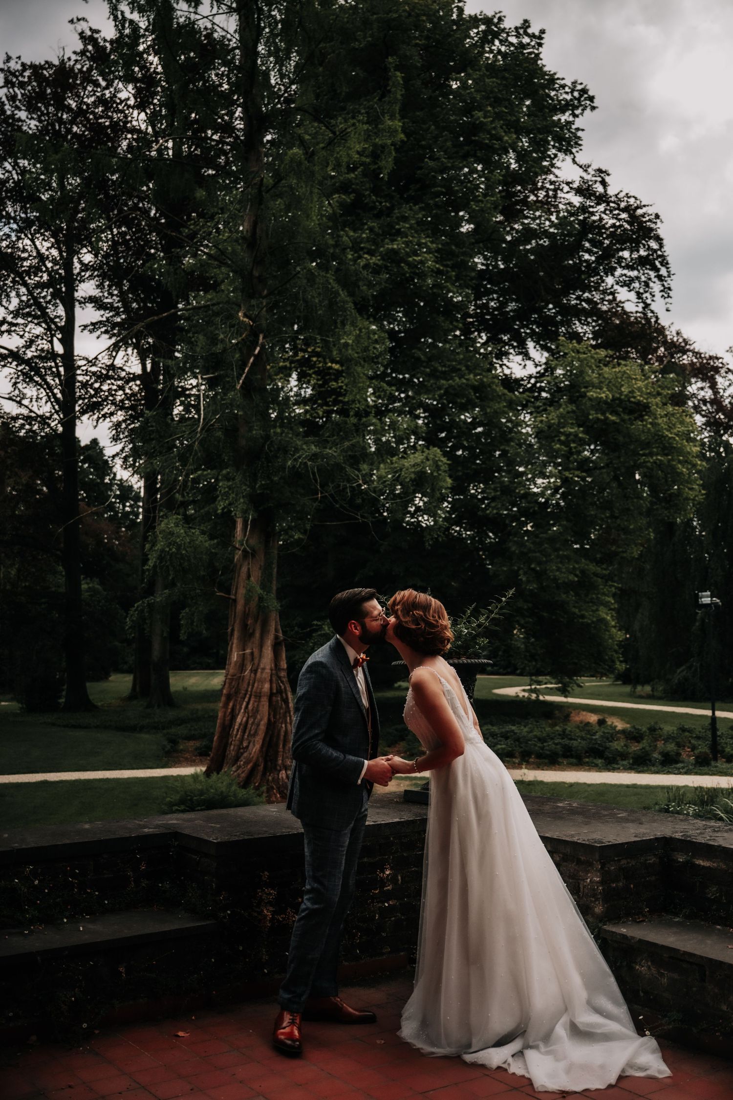 Couple sharing a tender moment on stone wall with lush trees in background.