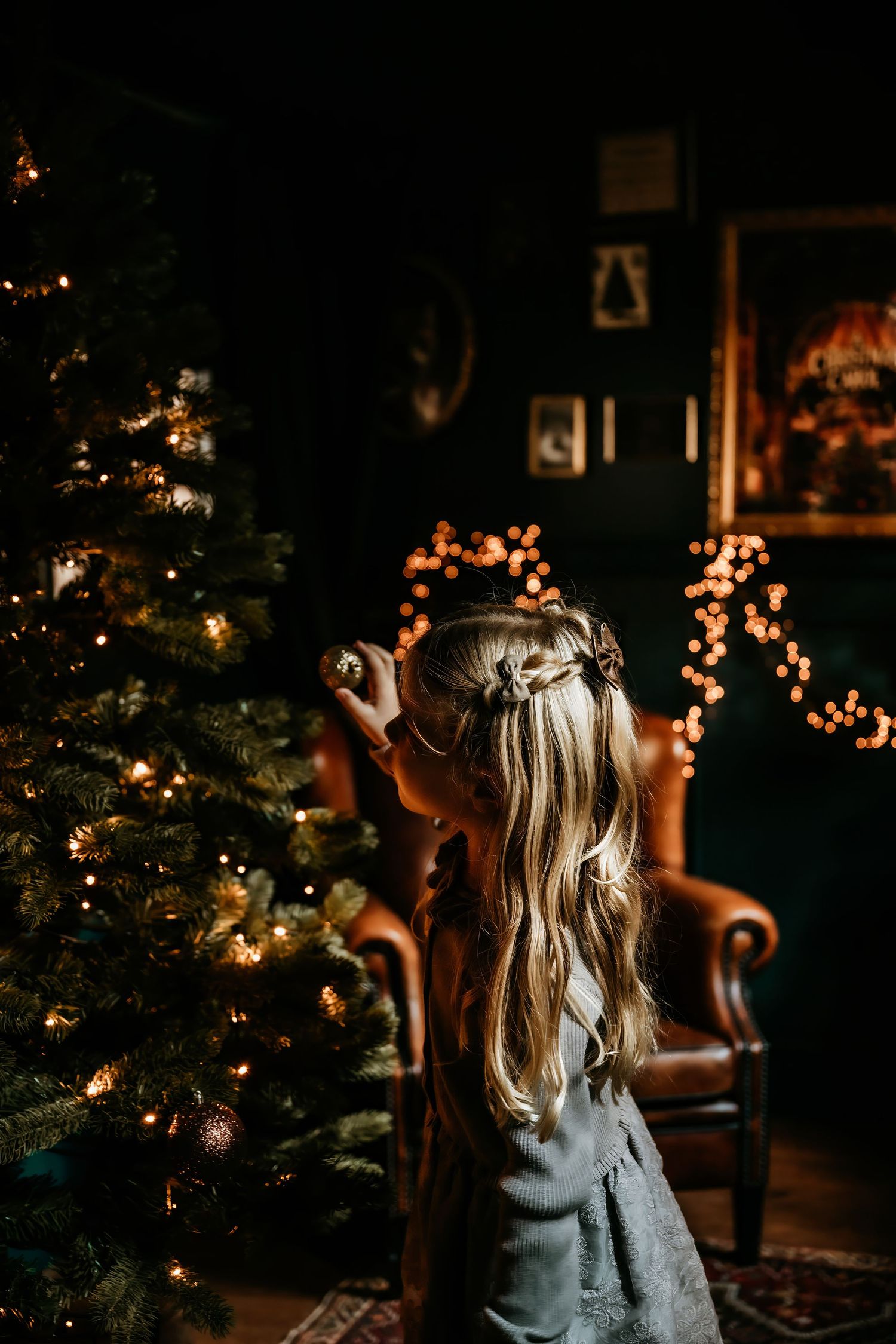 A child in a blue dress stands near a glowing Christmas tree in a dimly lit room.