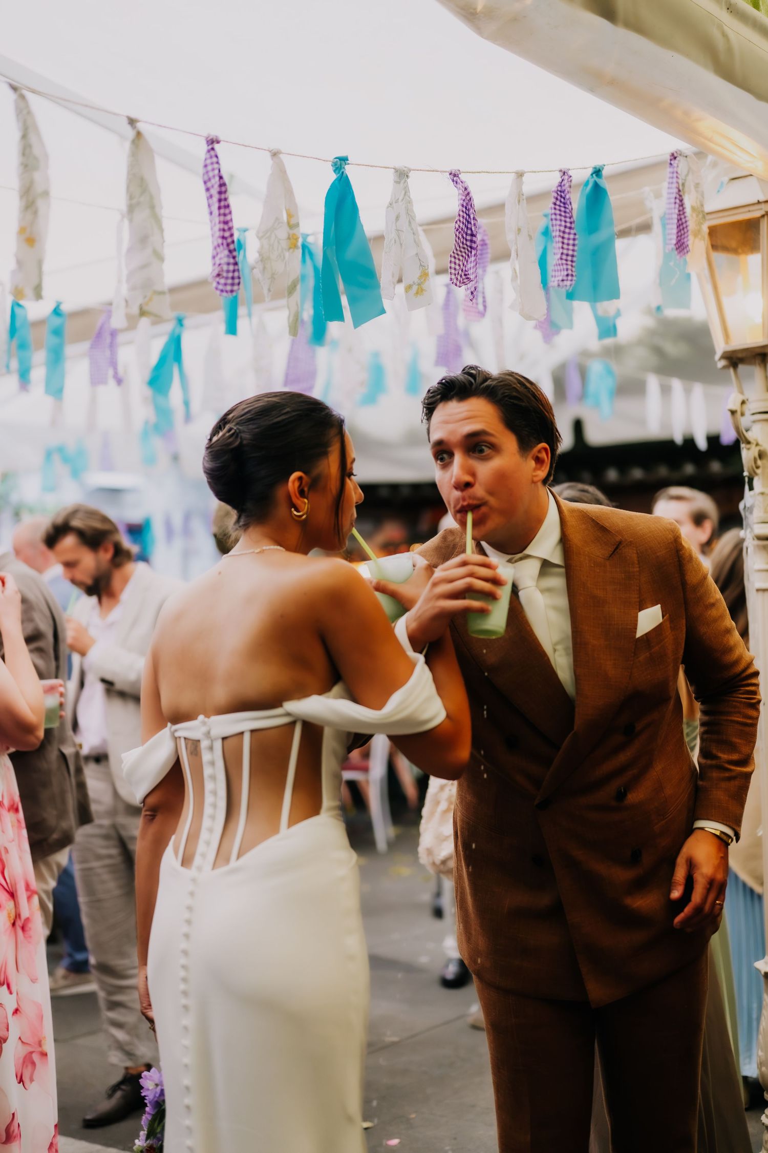 A bride in a white corset gown shares a drink with a groom in a brown suit at a festive outdoor wedding reception.