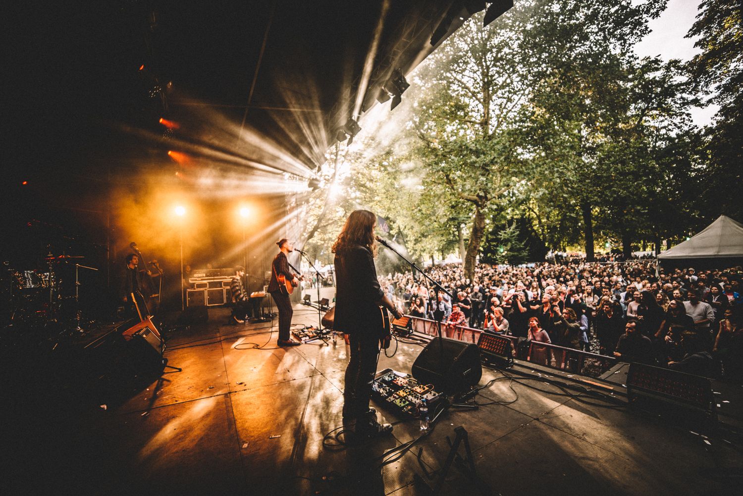 Silhouetted performers on stage at an outdoor music festival with dramatic sunset lighting and crowd in background.