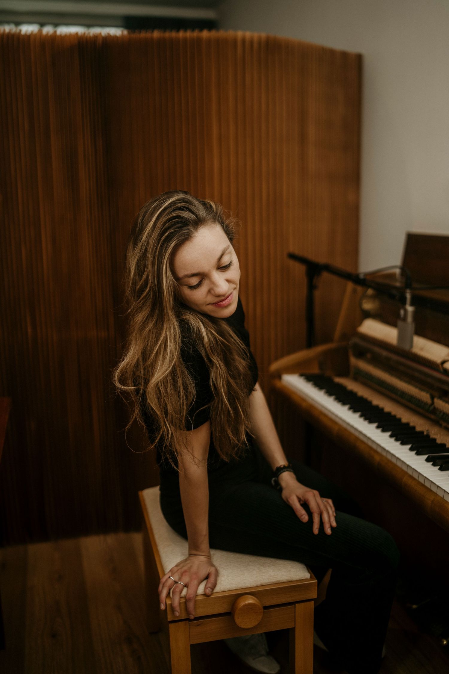 A woman in a black dress sits gracefully at a piano in a warmly lit room with wood paneled walls.