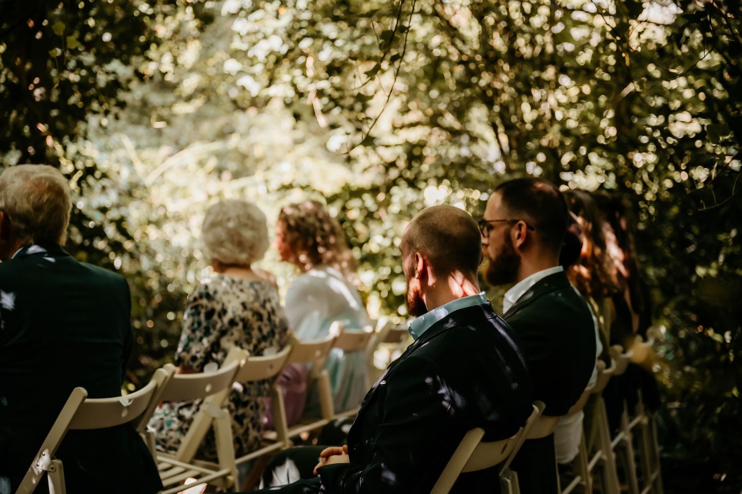 Wedding guests seated outdoors under natural lighting and tree canopy.