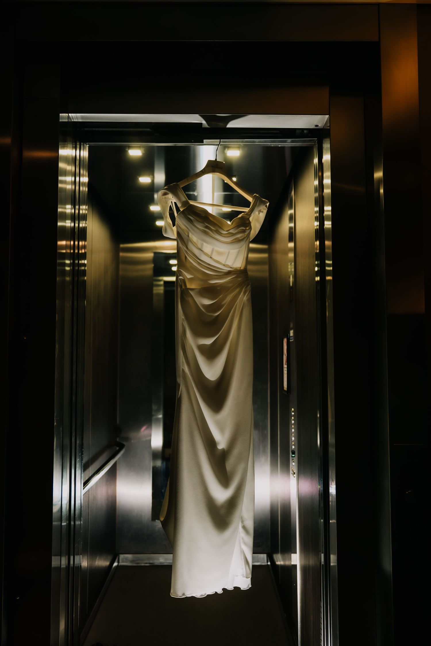 Elegant ivory satin wedding gown hanging on a wooden hanger inside a sleek mirrored elevator.