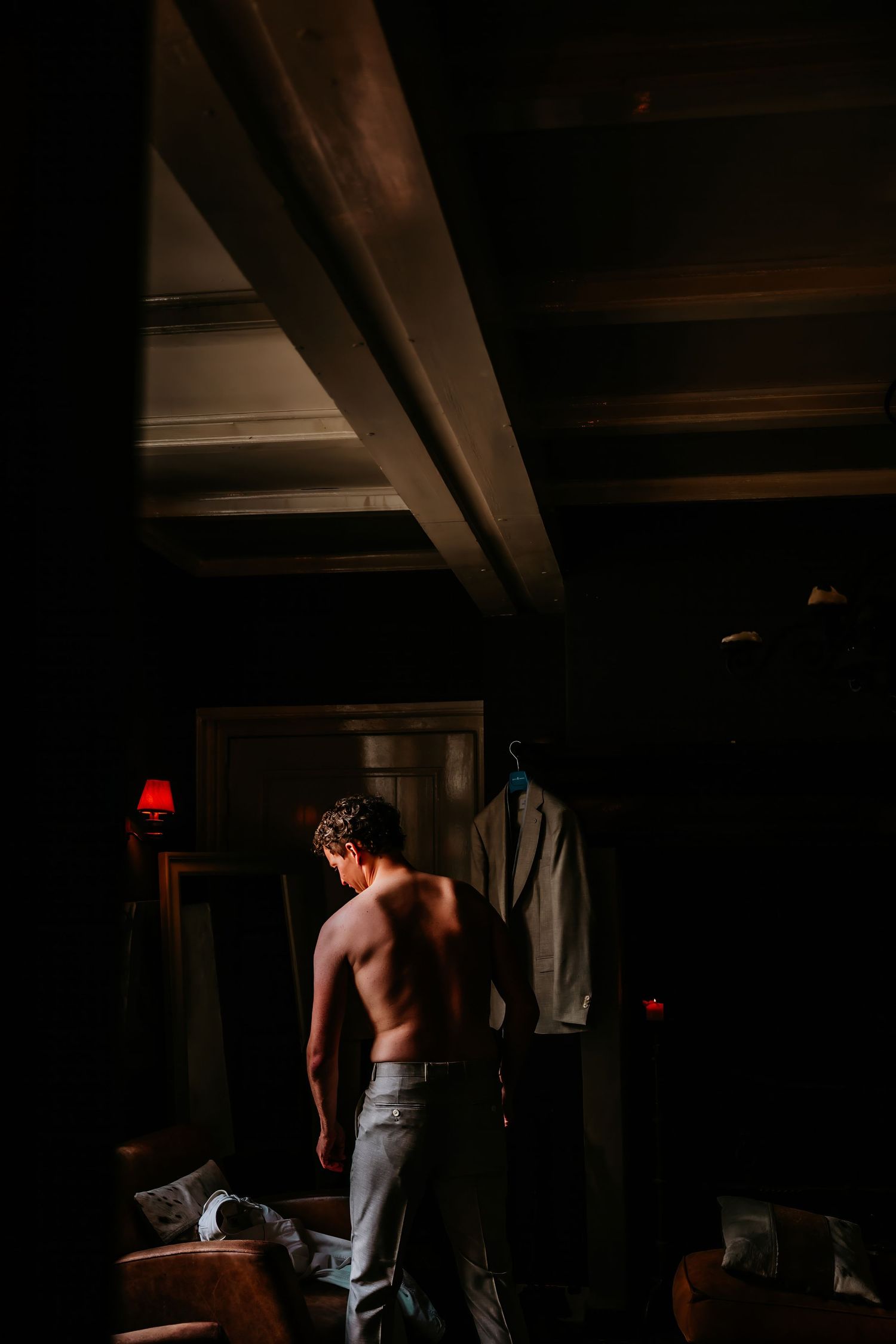 A shirtless figure stands in a dimly lit room with dramatic shadows and wooden beams visible overhead.