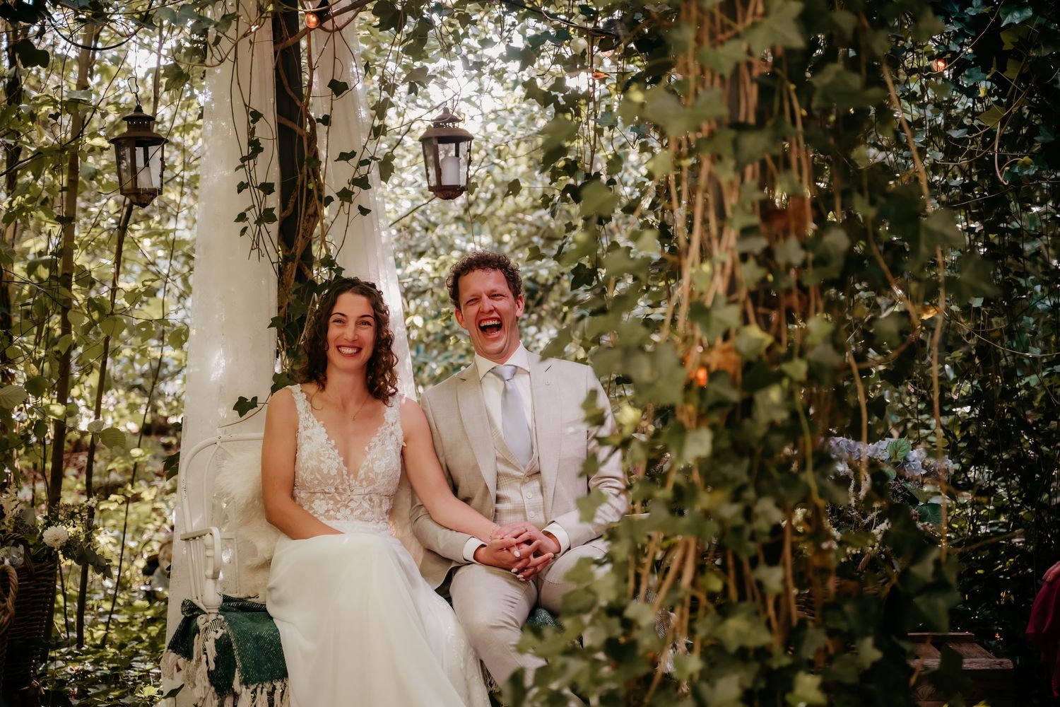 Newlyweds sit together on a garden bench surrounded by climbing vines and lanterns.