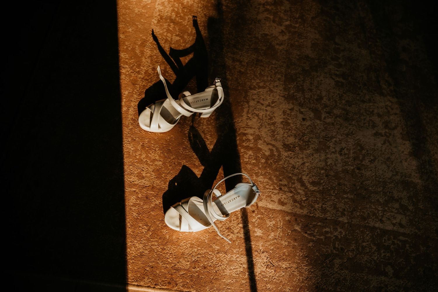 Wedding shoes casting dramatic shadows on a textured orange surface in artistic lighting.