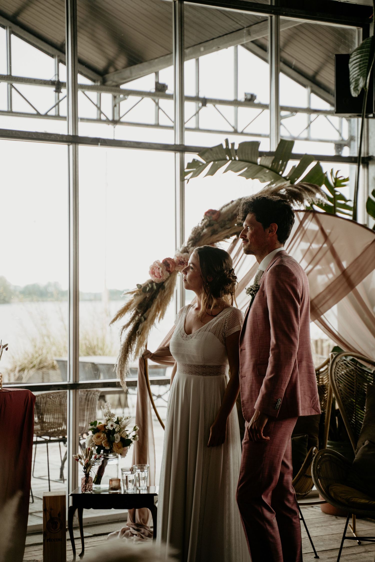 A couple stands together in a sunlit venue with large windows and tropical plants, creating an intimate wedding atmosphere.