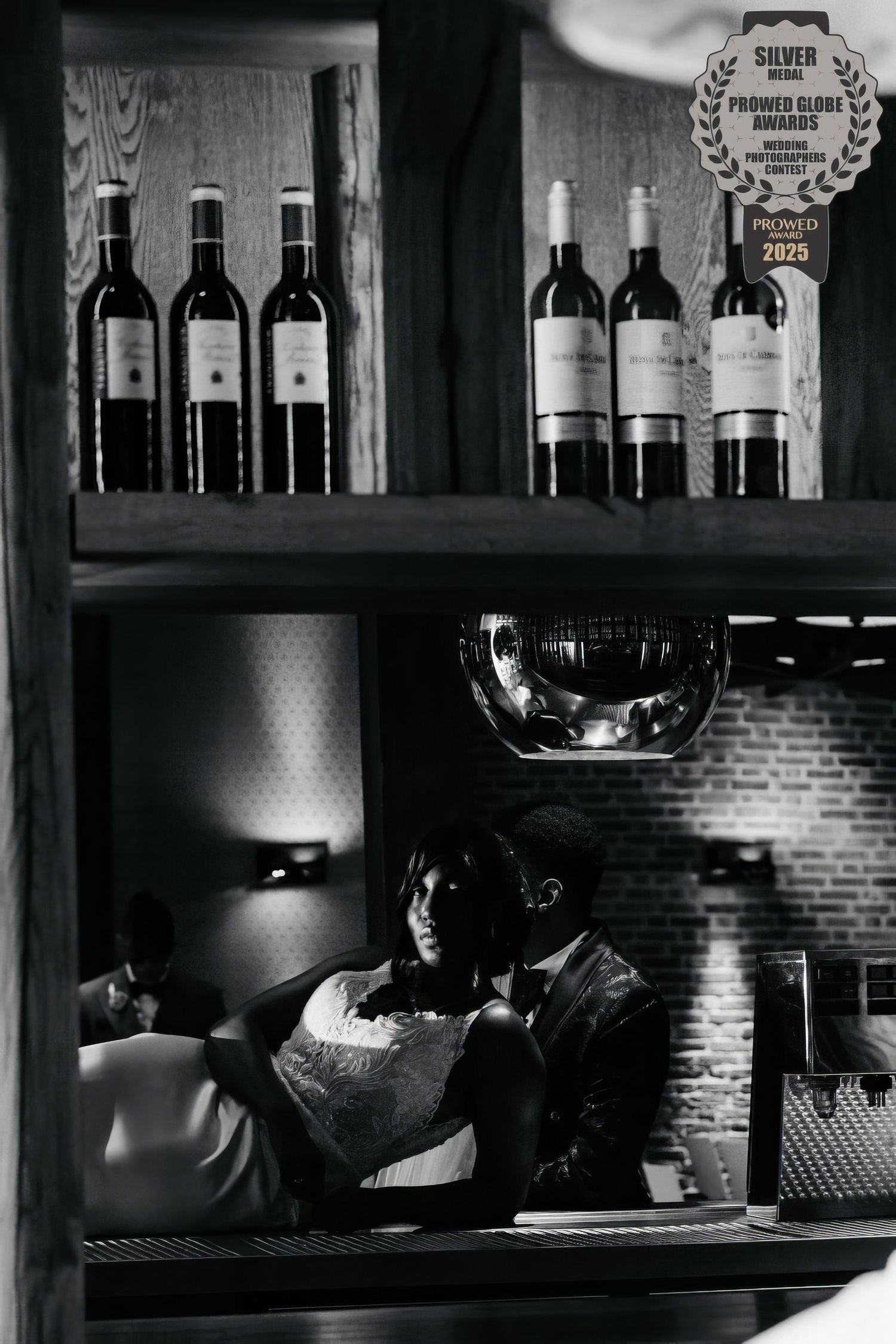 Black and white photo of a dimly lit bar interior with wine bottles displayed on shelves and brick walls.