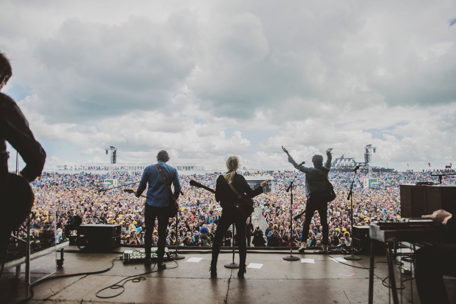 Silhouettes of performers on stage against a vast crowd at an outdoor music festival under cloudy skies.