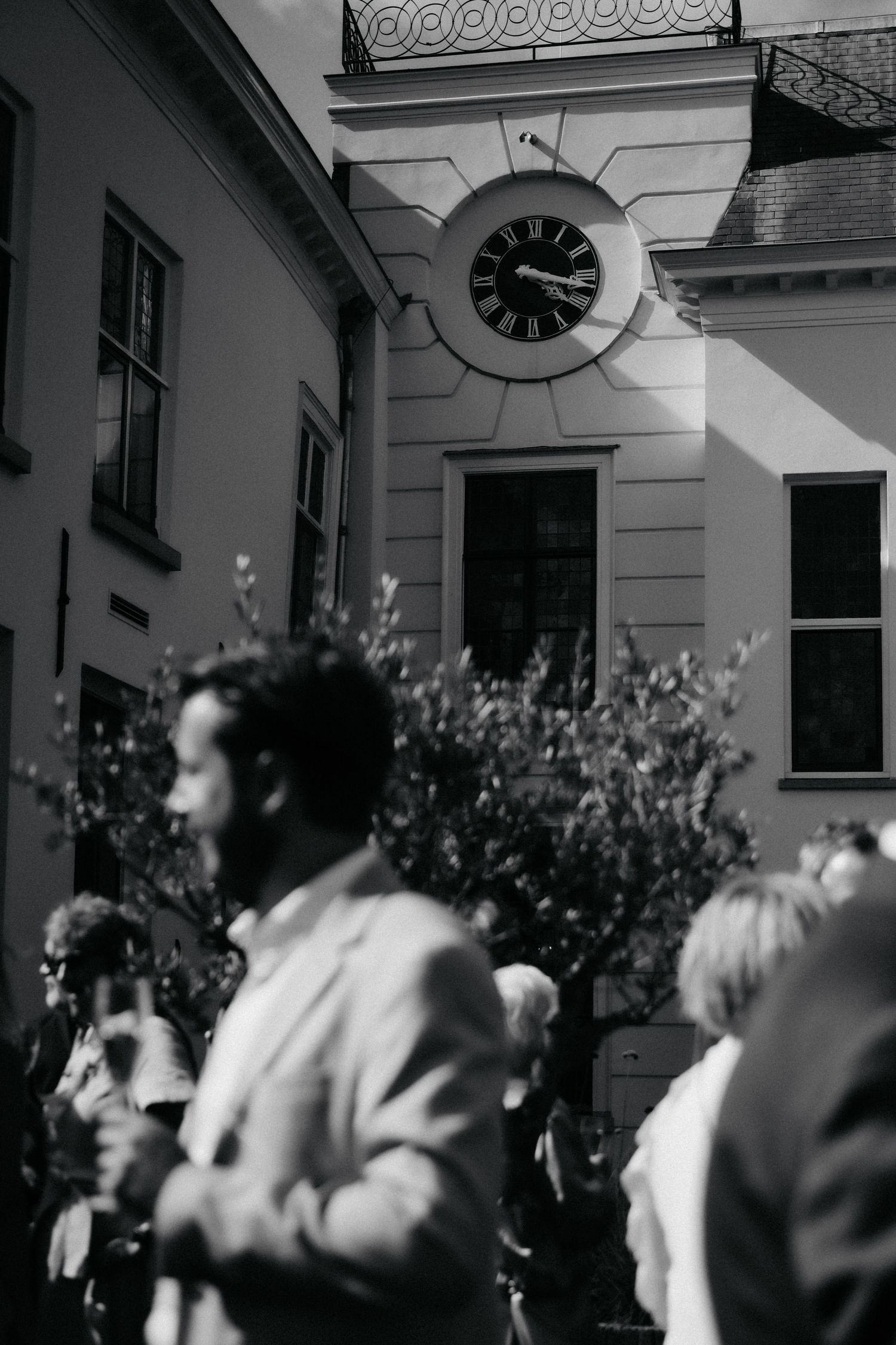 Black and white photo of elegantly dressed guests gathered outdoors near a classic building with a Roman numeral clock.