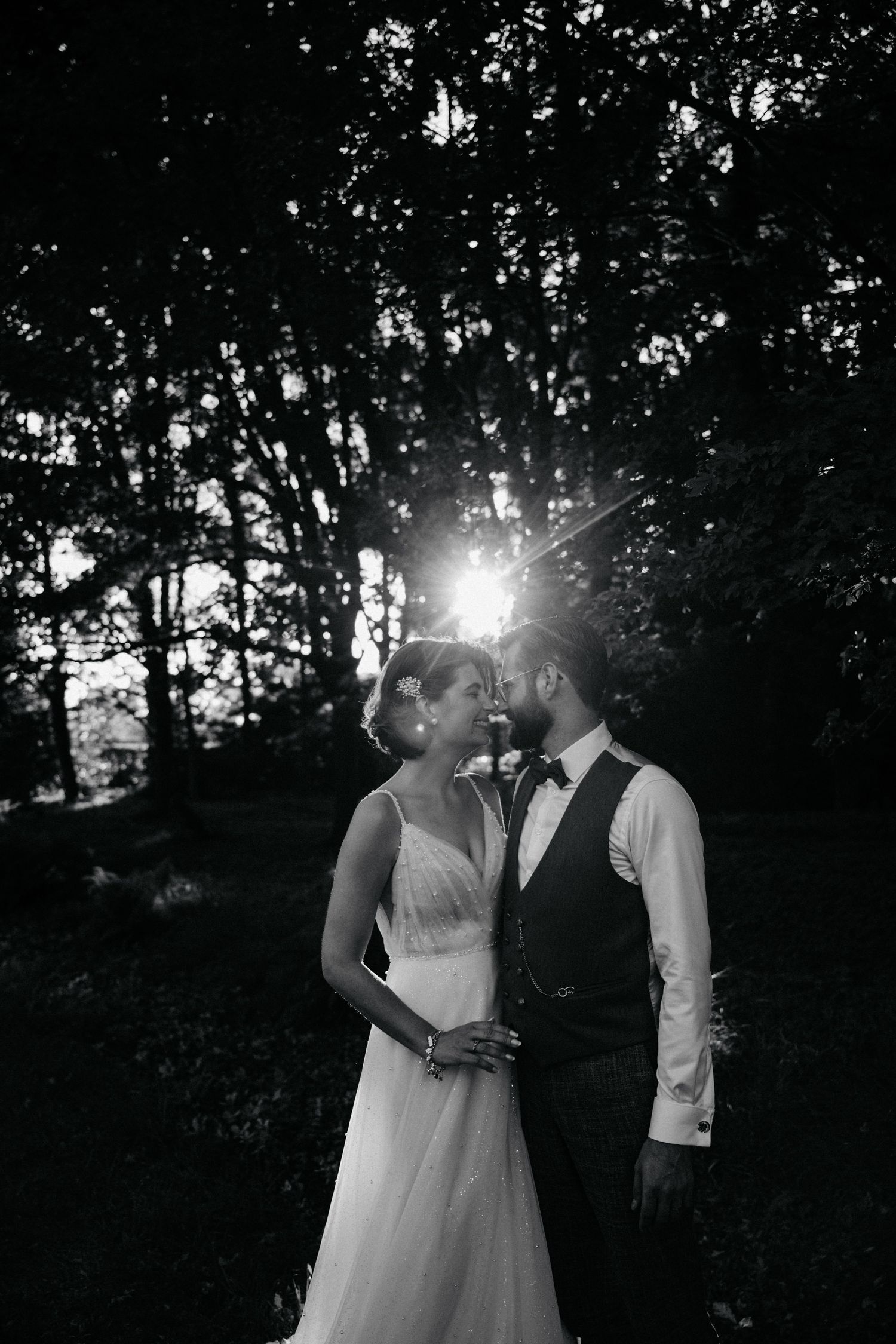 Black and white photo of a couple embracing during sunset with trees silhouetted behind them.