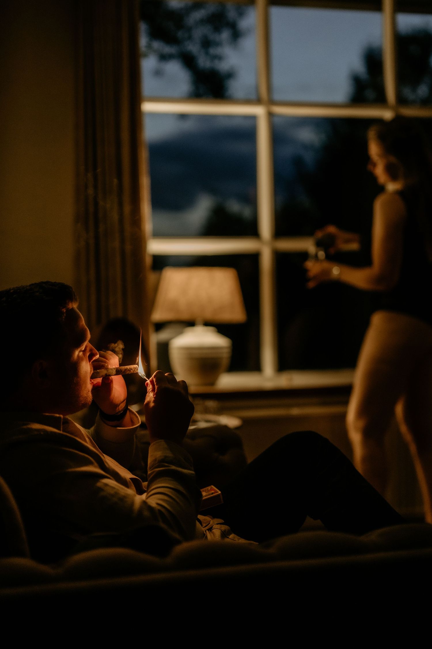 A cozy evening scene in dim lighting with people relaxing by a window with a table lamp casting warm ambiance.