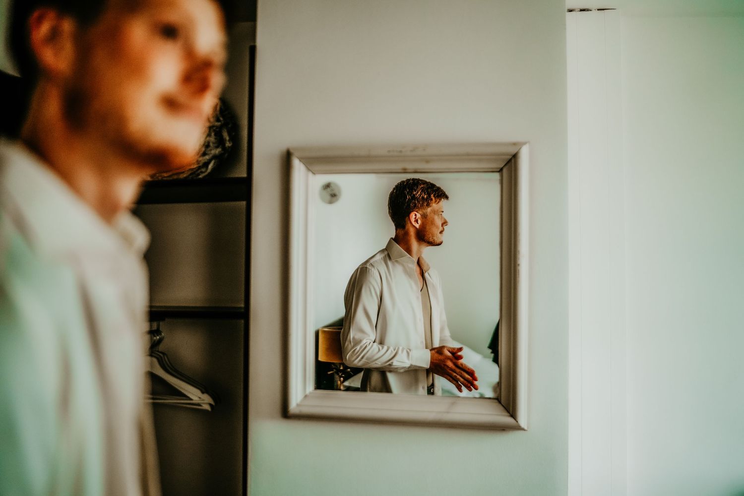 A person in white clothing stands near a window frame, creating a minimalist composition with soft natural lighting.