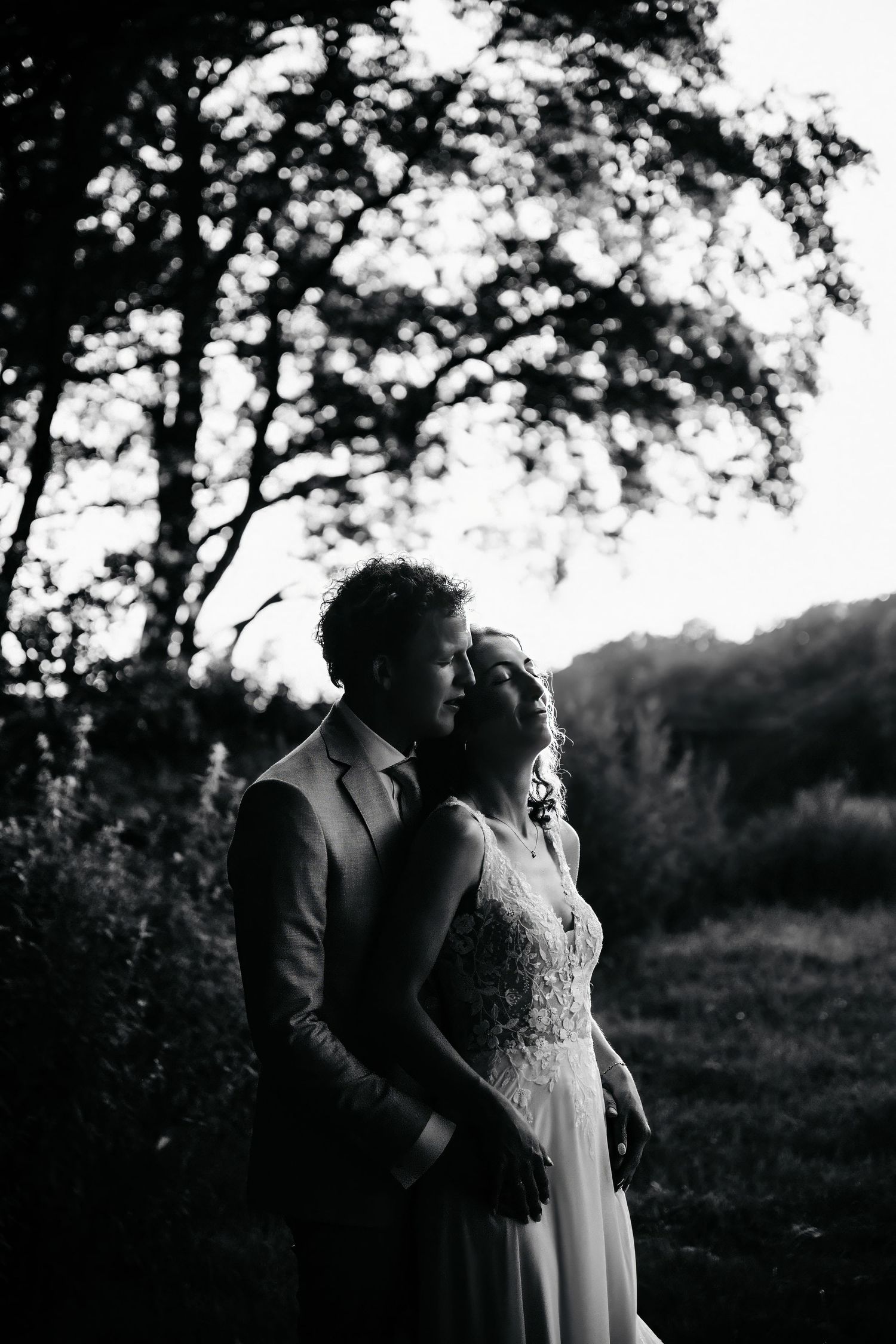 Romantic black and white portrait of wedding couple under trees at sunset.