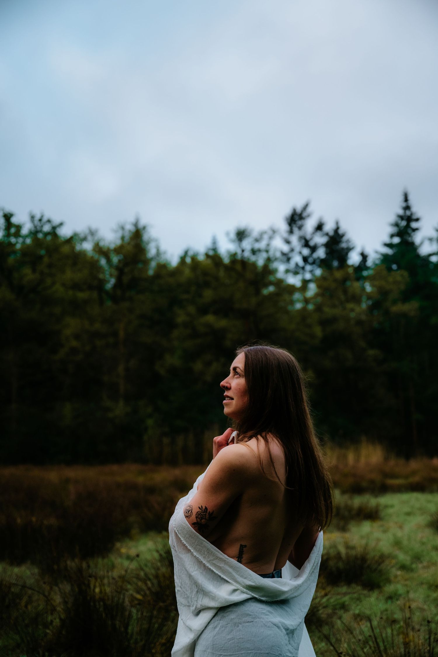 Woman in white sweater poses in nature against backdrop of evergreen trees at dusk.
