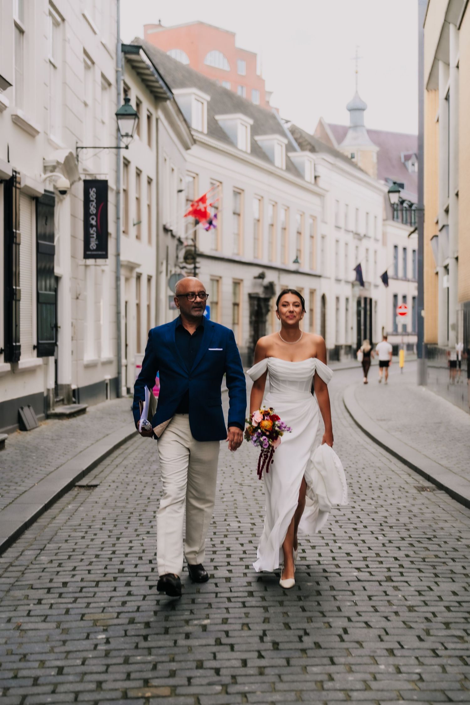 Bride and groom walking hand-in-hand on cobblestone street in European city, bride holding colorful bouquet in white off-shoulder gown.