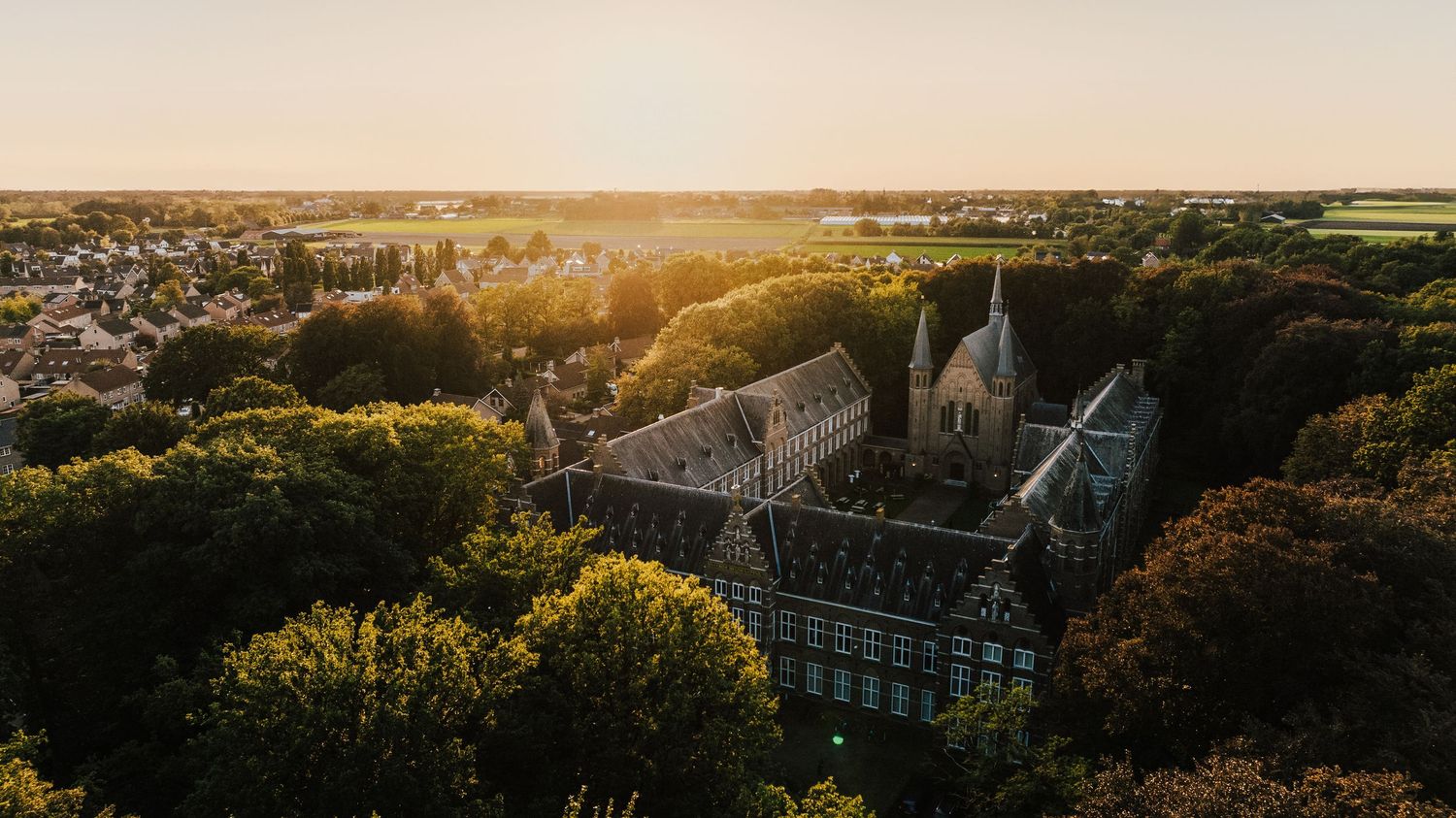 Aerial view of a Gothic cathedral surrounded by autumn trees at sunset with golden light streaming across the landscape.