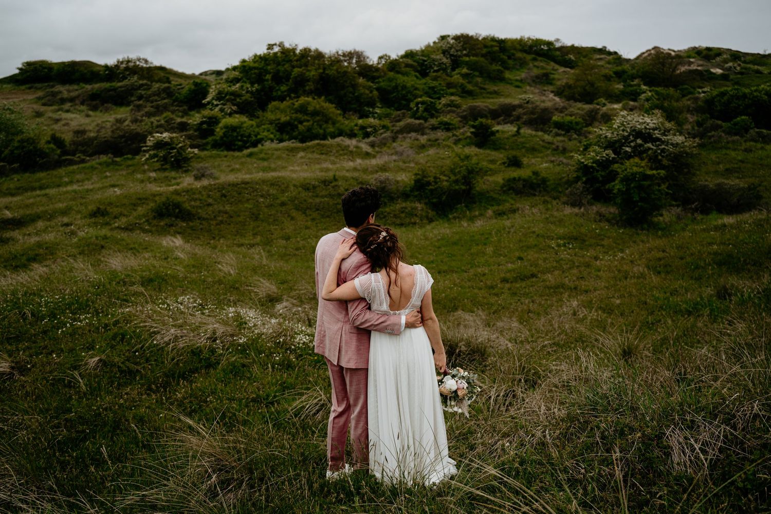 Wedding couple embracing on a scenic hilltop with green vegetation and overcast sky.