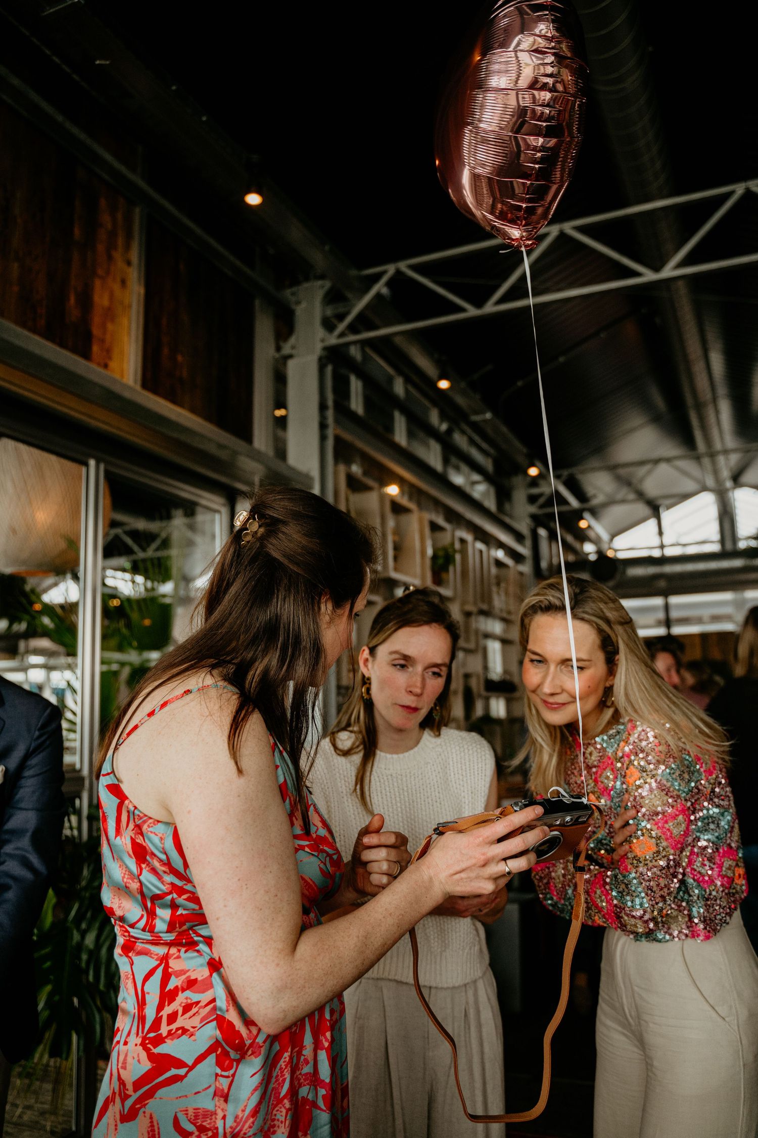 Three friends looking at a phone together under a balloon at an indoor event.