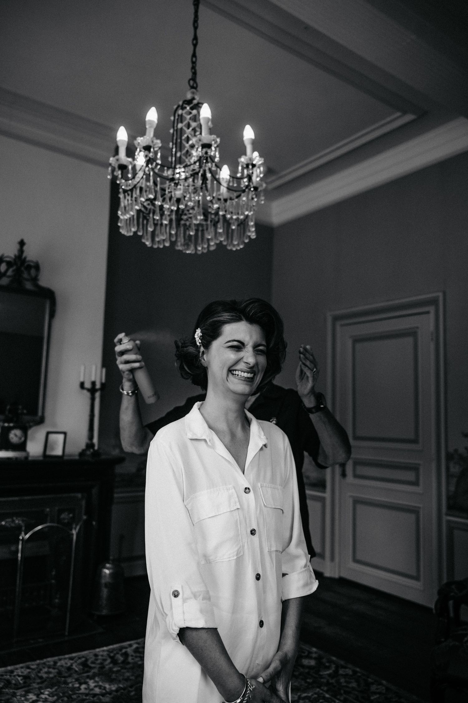 A person in a white button-down shirt laughs joyfully under a crystal chandelier in an elegant room.