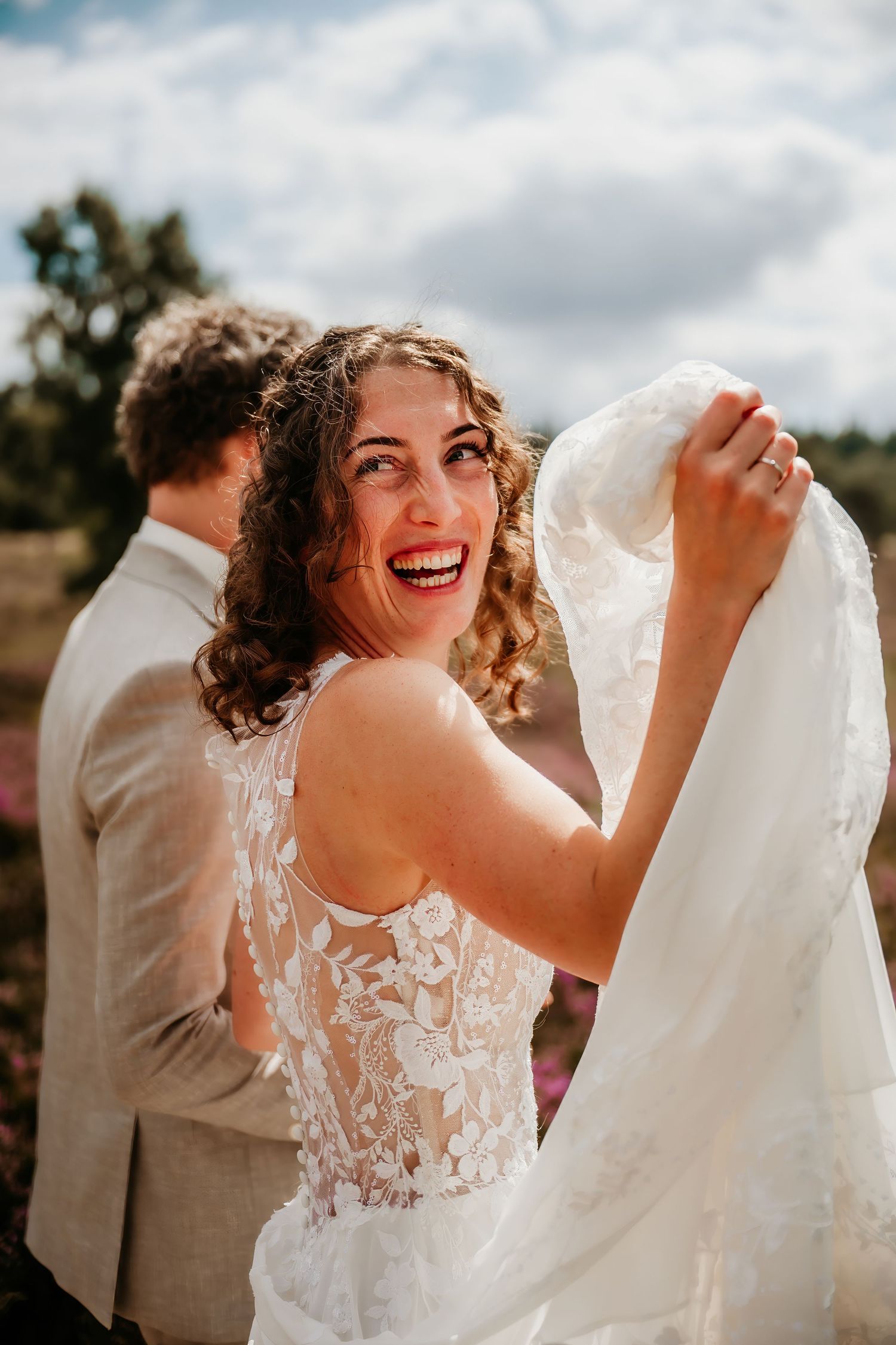 A joyful bride in a lace wedding dress laughs while holding her veil outdoors on a sunny day.