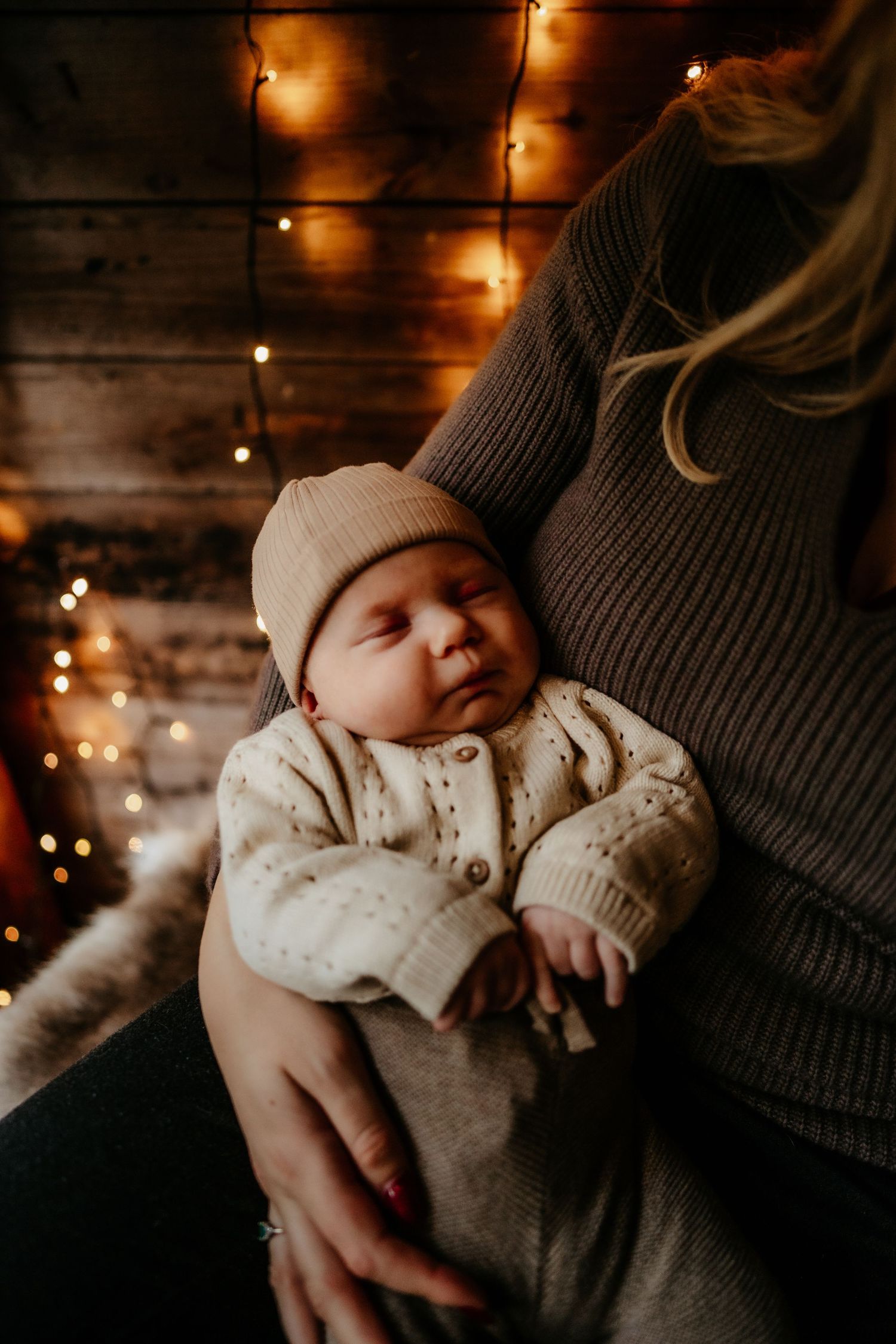 A sleeping baby in a knit sweater and beanie is cradled against someone wearing a gray sweater, with warm twinkle lights behind.
