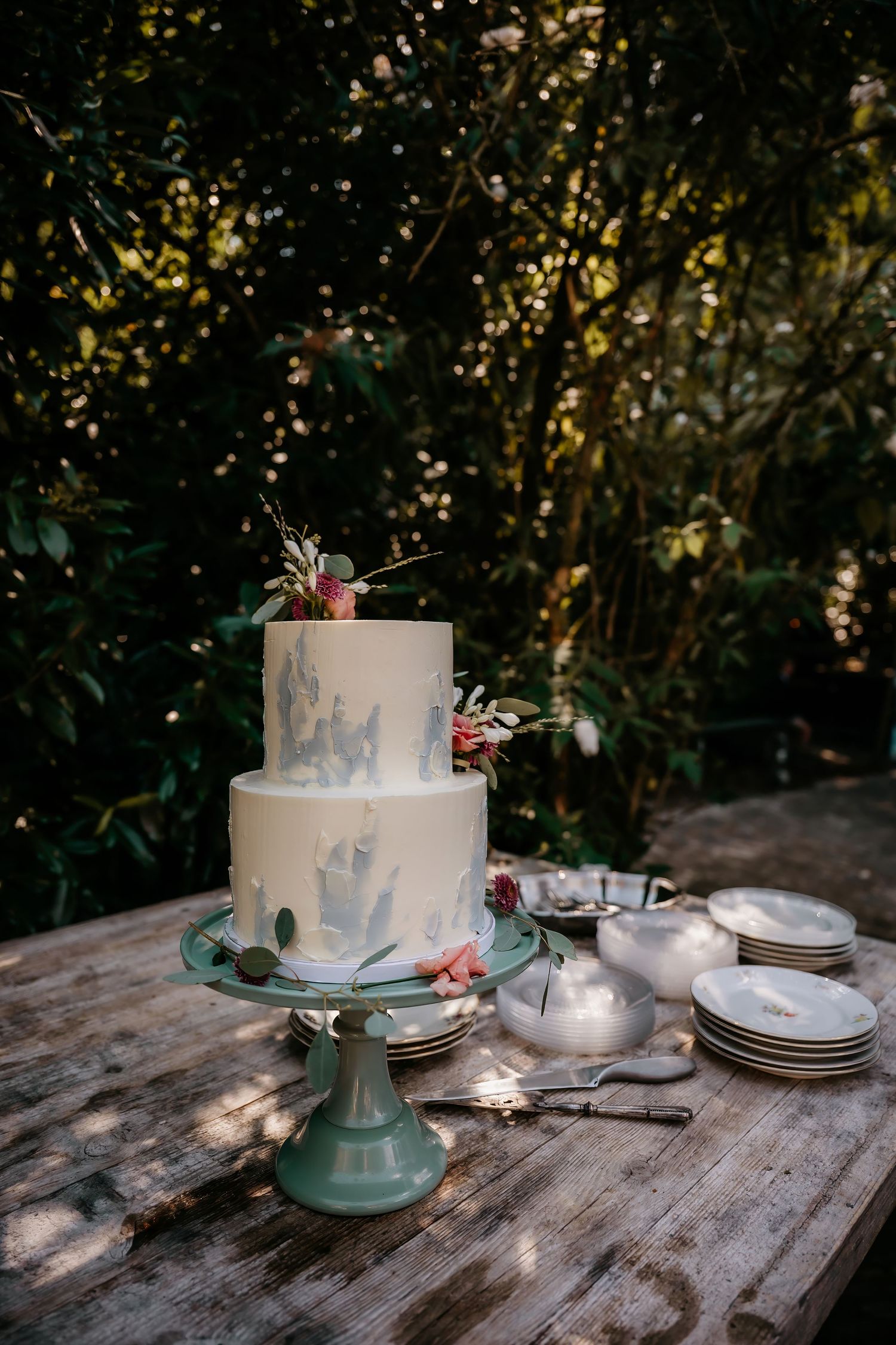 An elegant two-tier marble-effect wedding cake displayed on a turquoise stand in dappled sunlight.