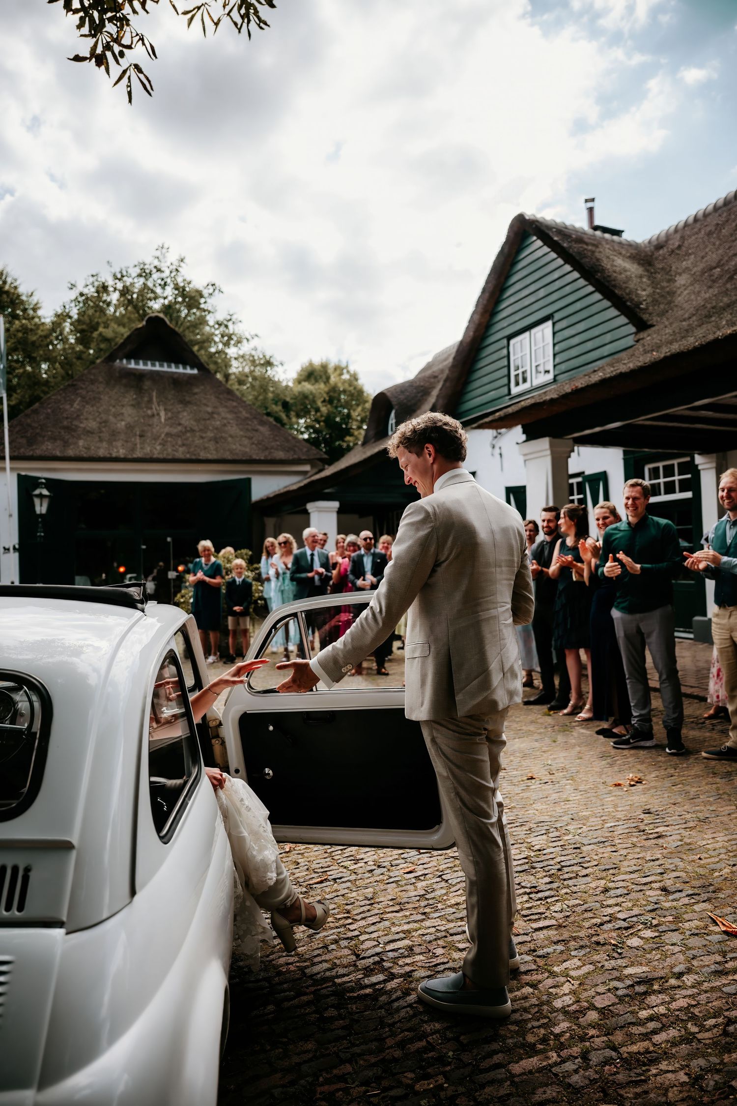 A classic vintage Volkswagen Beetle parked outside a rustic thatched-roof cottage during a wedding celebration.