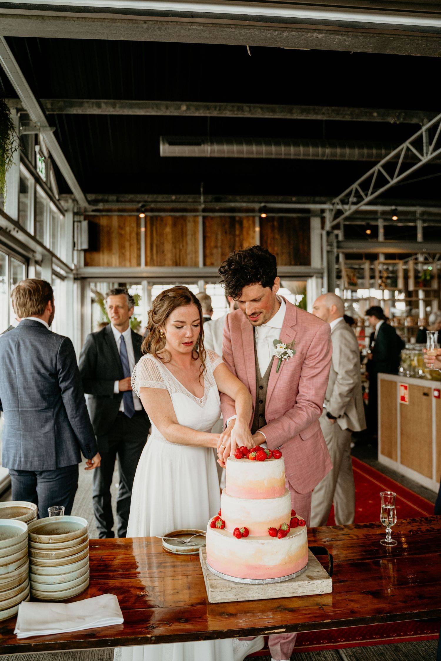 A couple cuts their three-tiered white wedding cake decorated with red berries in an industrial venue.