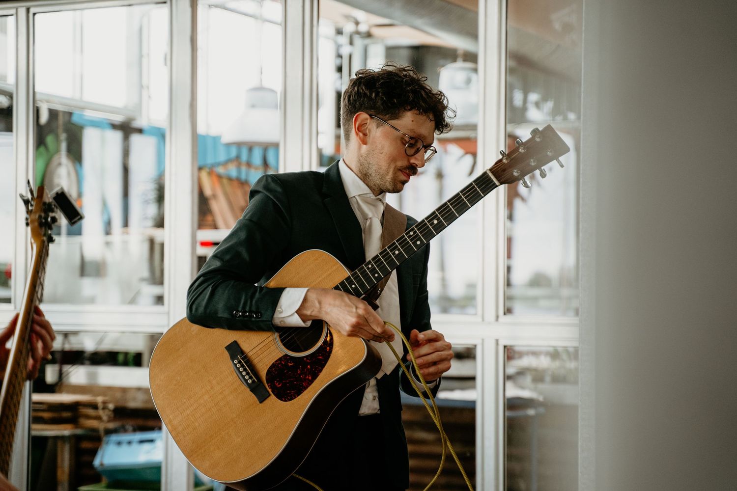 A musician plays acoustic guitar by large windows in natural light.
