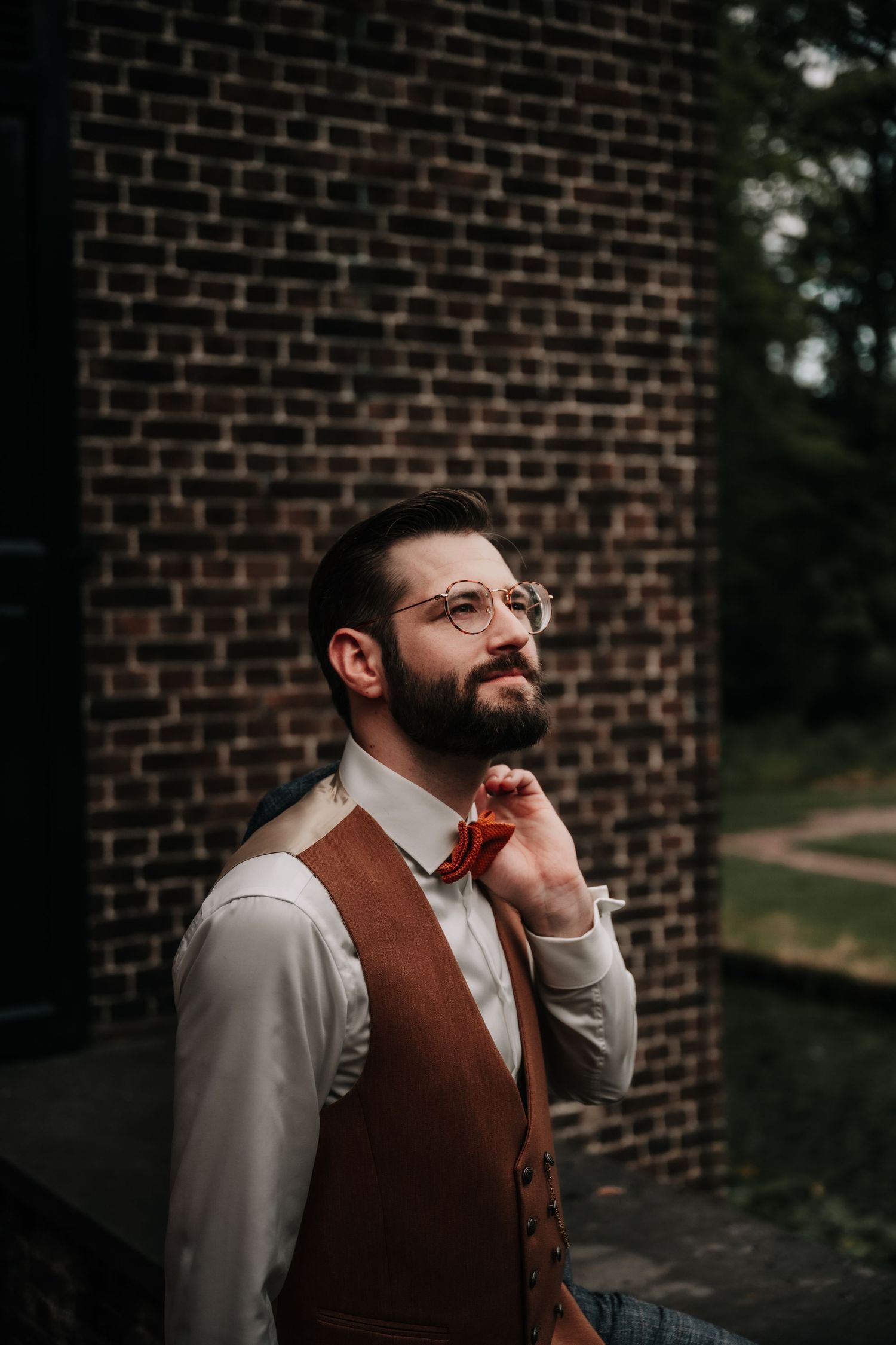 Person in brown vest and glasses against brick wall in moody lighting.