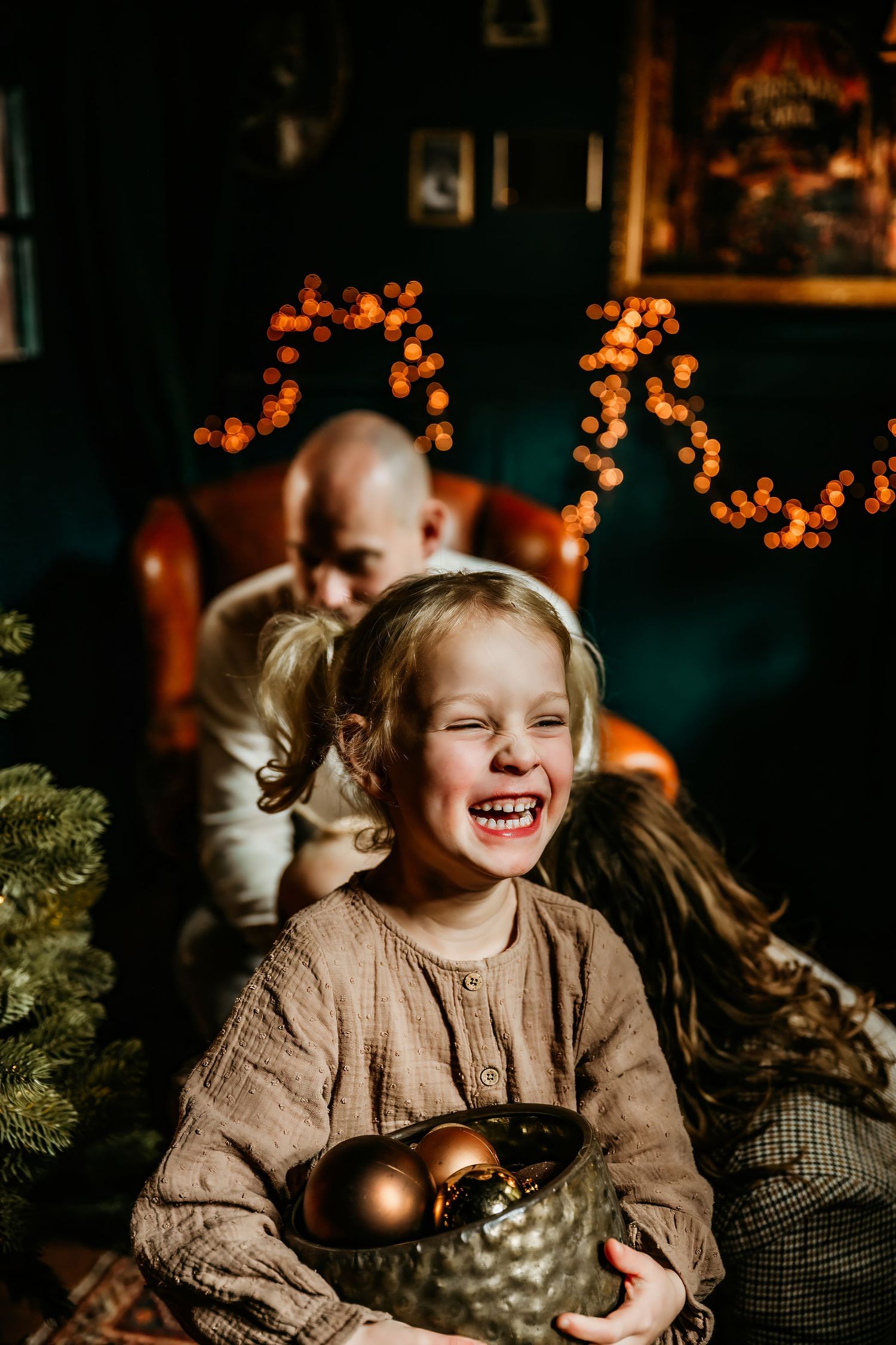 A child in a brown sweater laughs joyfully while sitting near Christmas decorations and twinkling lights.