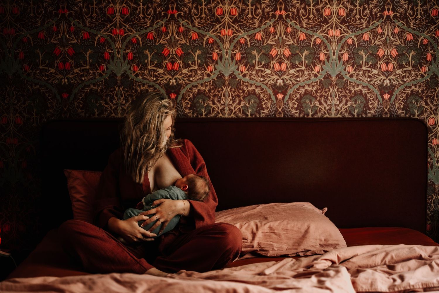 A tender moment in a dimly lit bedroom with ornate red wallpaper and burgundy bedding.