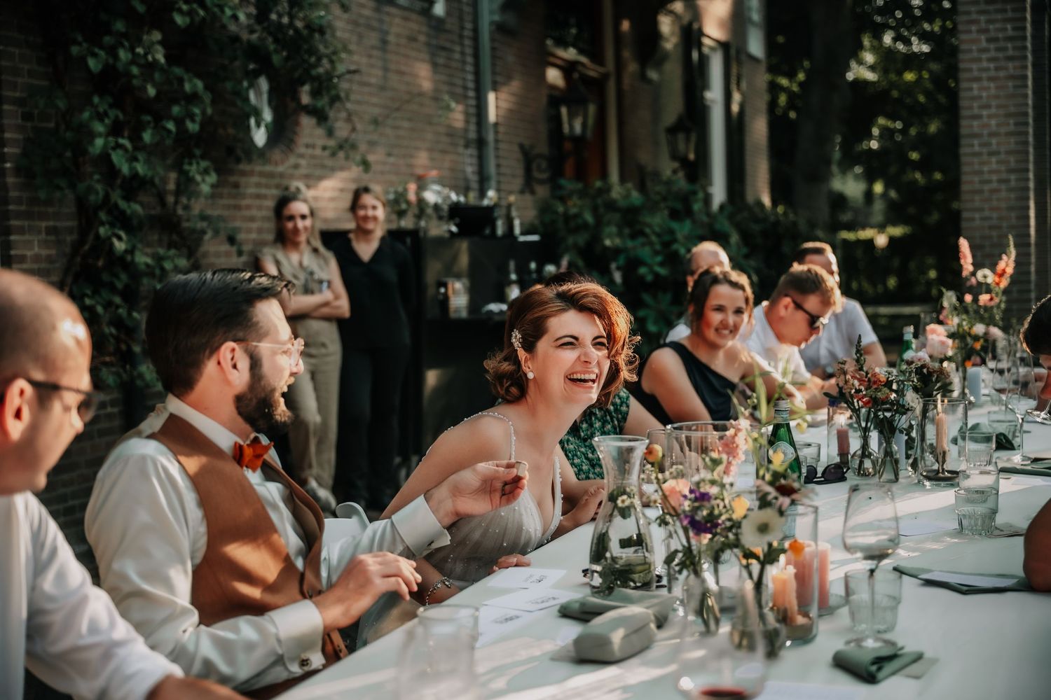 Wedding reception guests laugh and smile while seated at an elegant outdoor dining table with floral centerpieces.