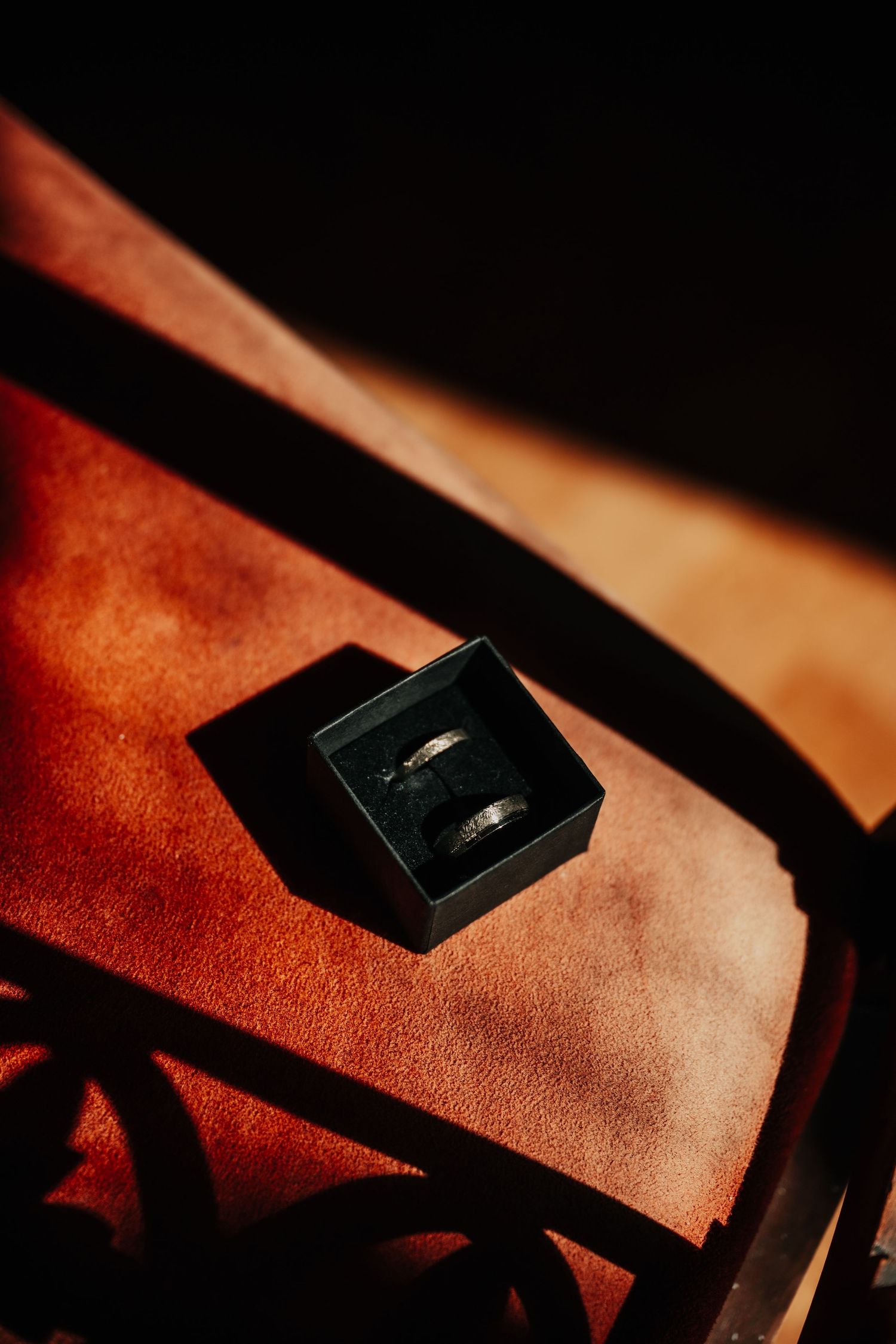 A wedding ring sits in a small black box casting dramatic shadows on a leather surface.
