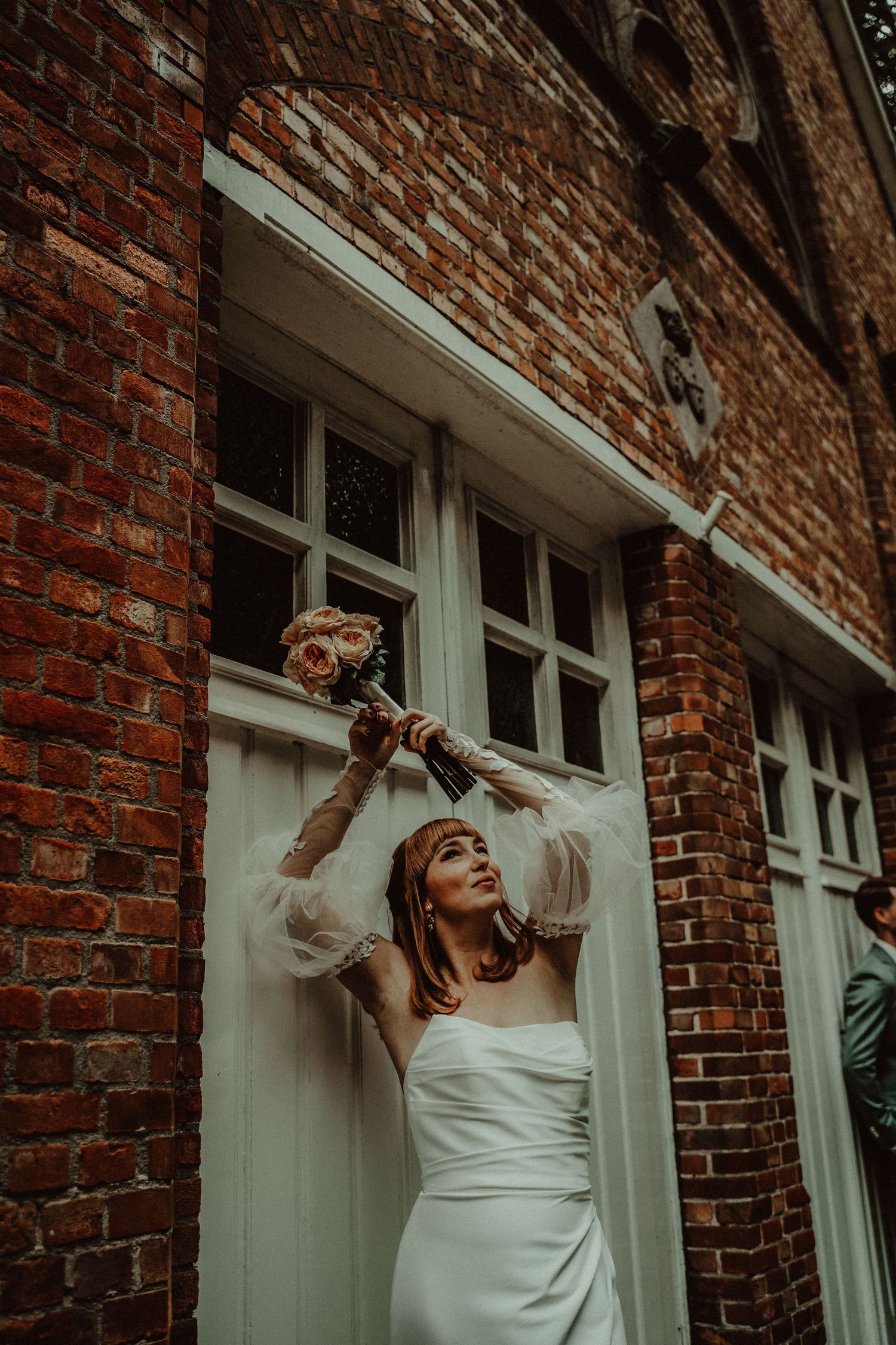 A bride in a white dress poses playfully against a rustic brick wall with white-framed windows.