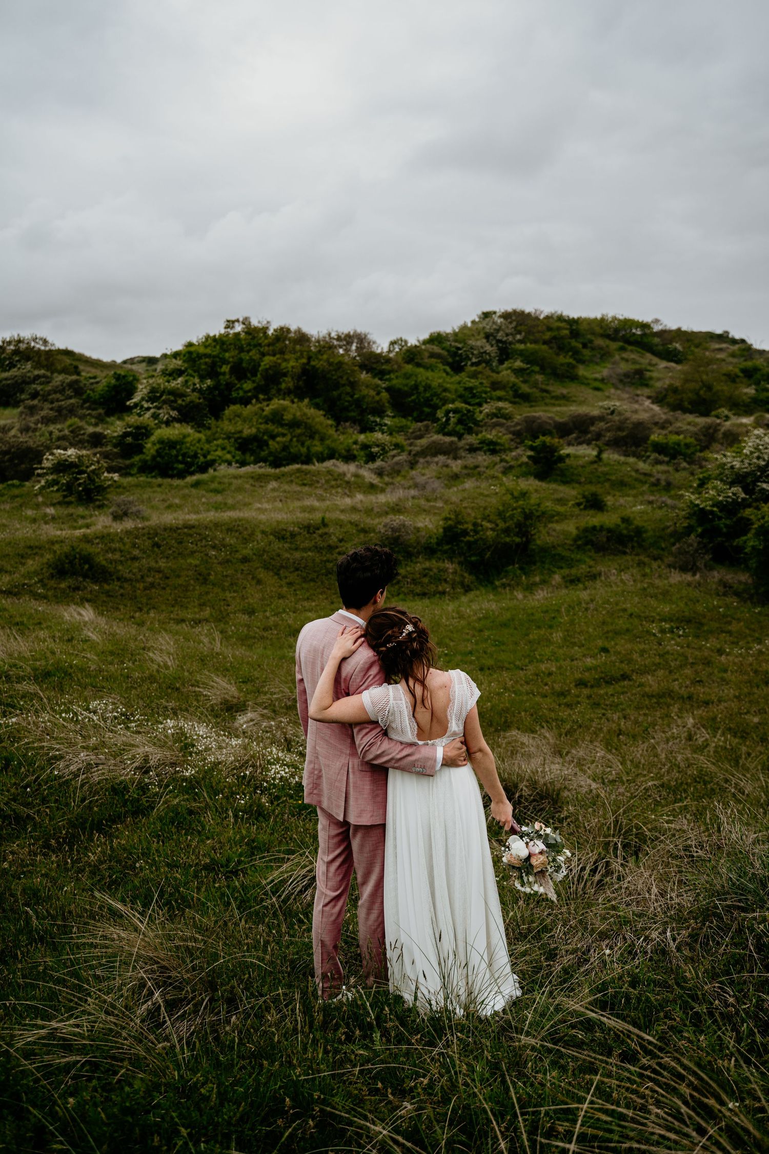 Couple in wedding attire walking together on a grassy hillside under cloudy skies.