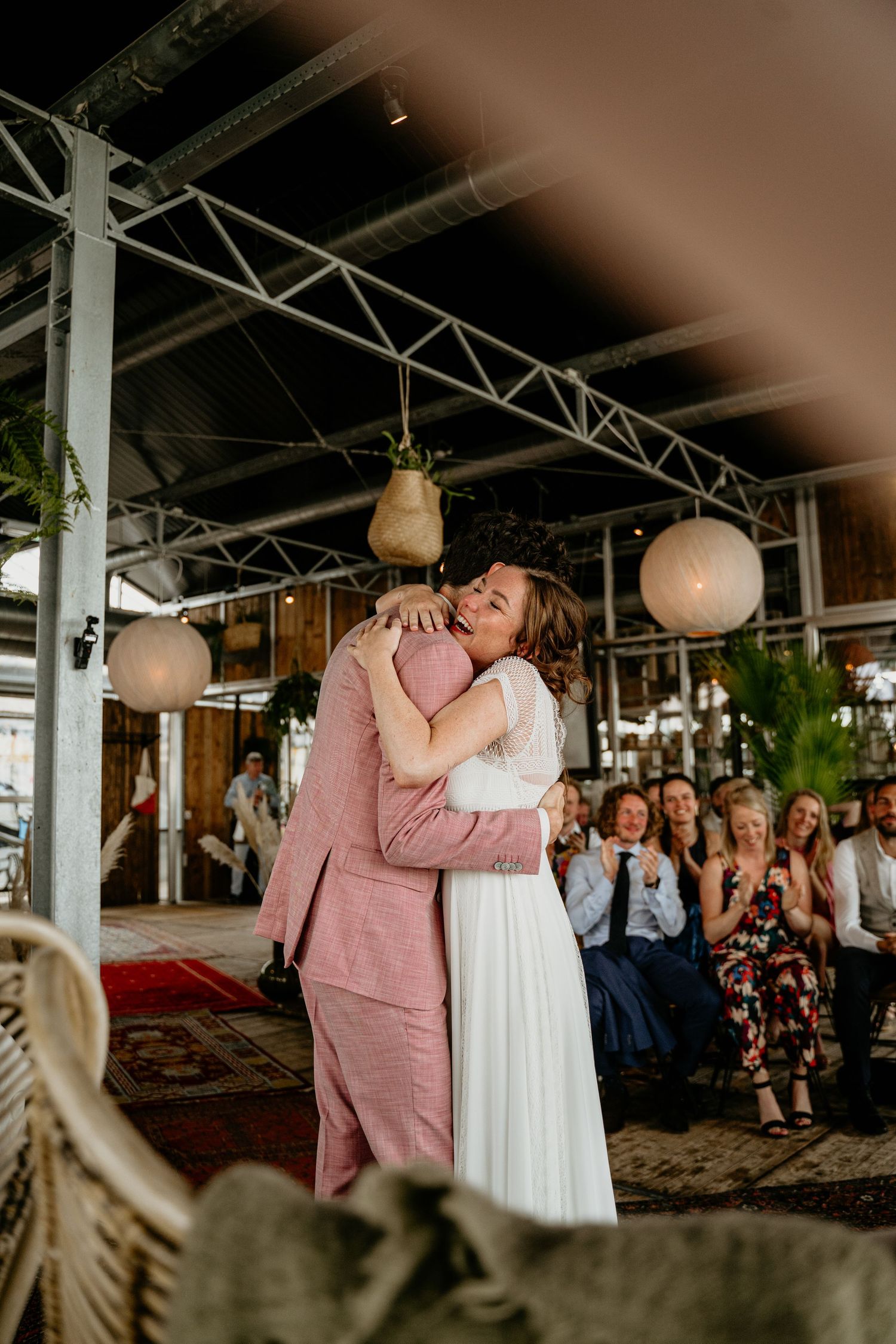 A couple embraces during a celebration under an industrial-style venue ceiling with hanging lanterns.