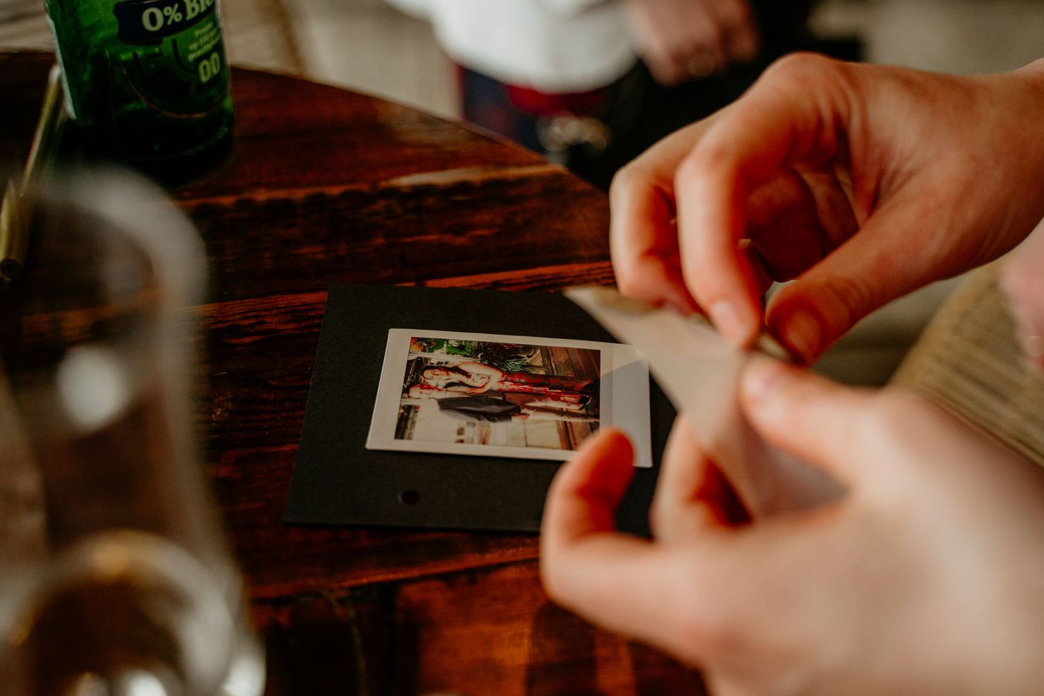 Hands hold a polaroid photograph on a dark wooden table with drinks in the background.