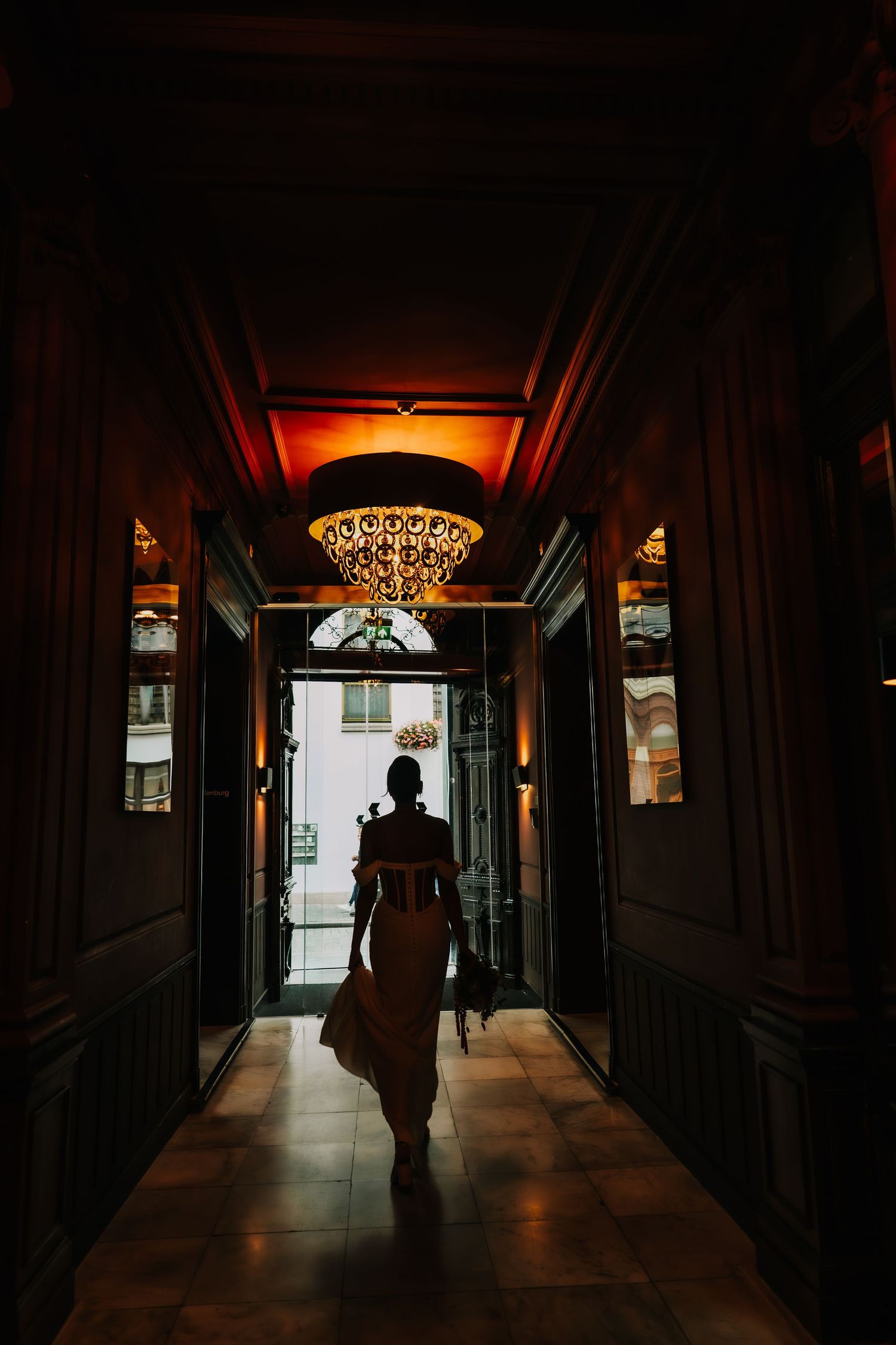 Silhouette of a woman in a elegant dress walking through a dimly lit hallway with a chandelier.