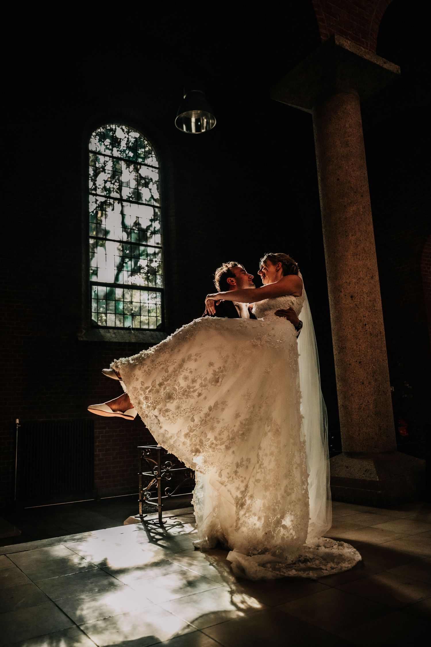 A bride in a flowing white dress twirls elegantly in a dark church interior illuminated by stained glass windows.