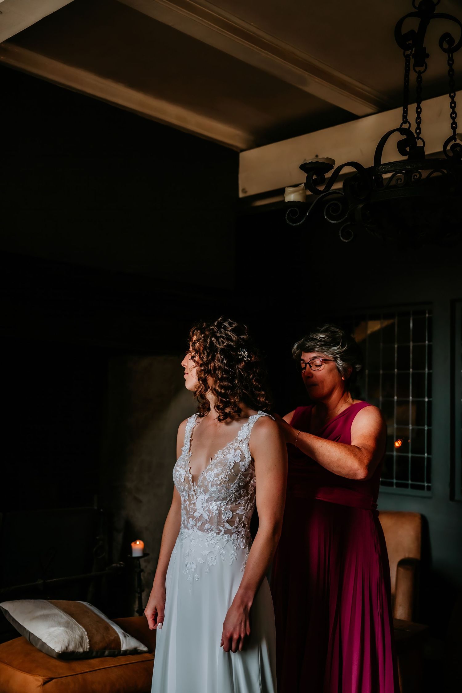 A wedding dress fitting moment captured in moody lighting beneath a decorative chandelier.