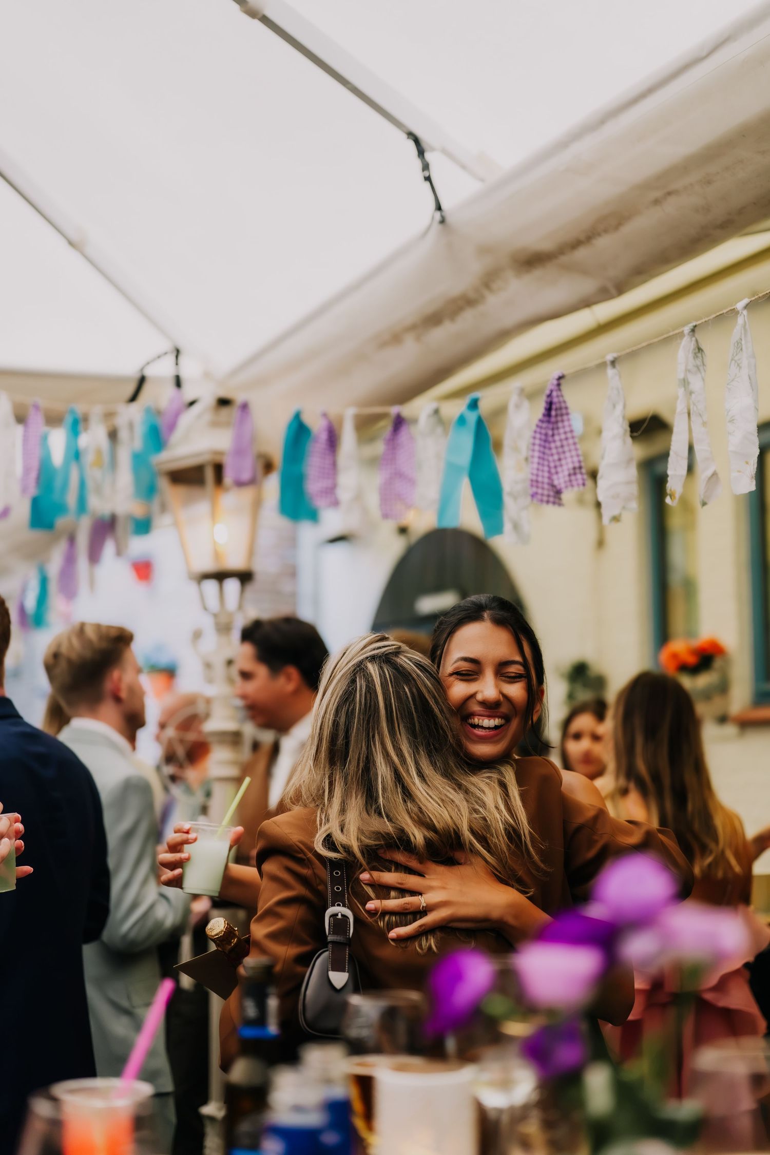 Two women share a warm hug at a festive outdoor party with colorful bunting decorations overhead.