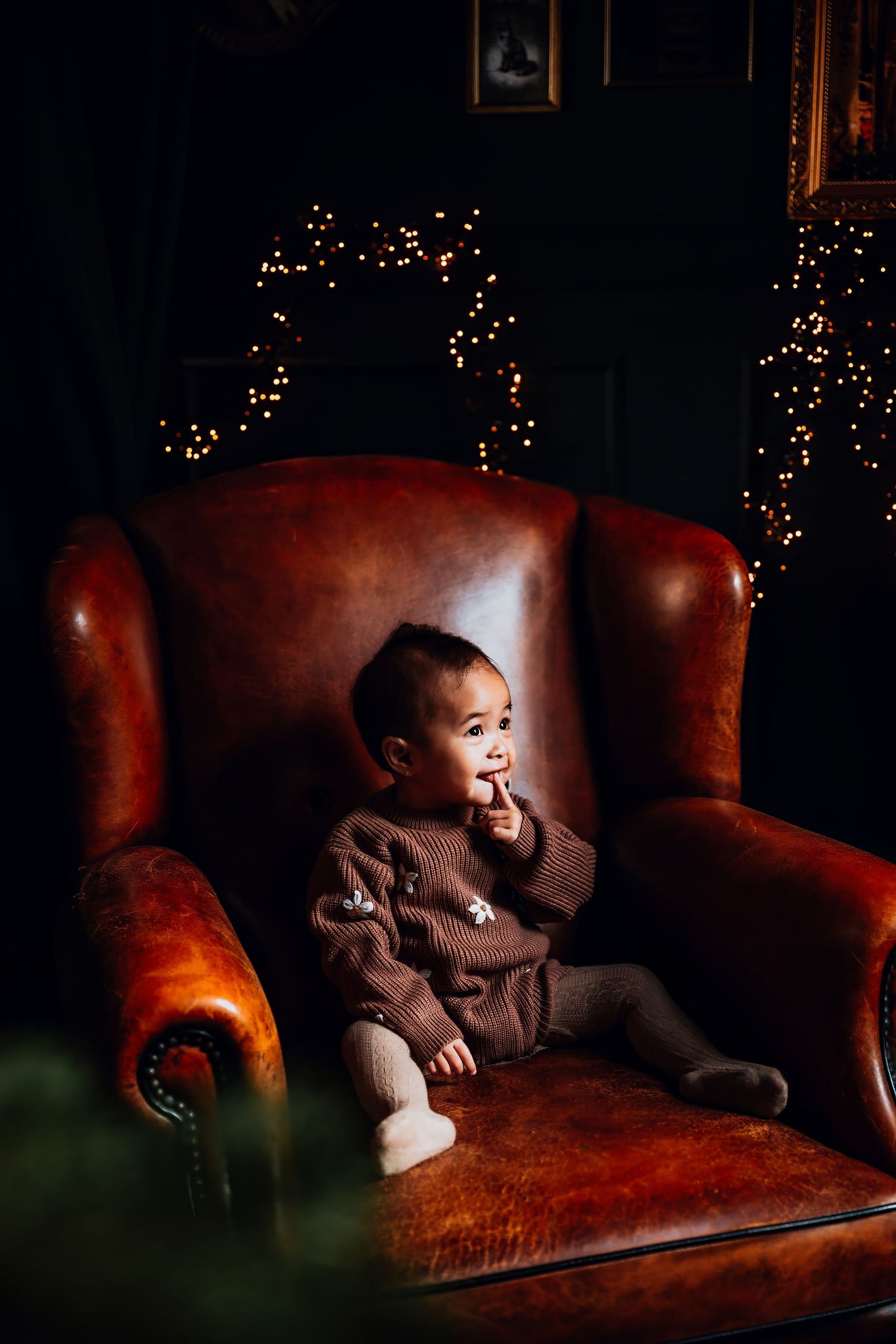 A toddler sits in a large leather armchair with twinkling Christmas lights in the background.
