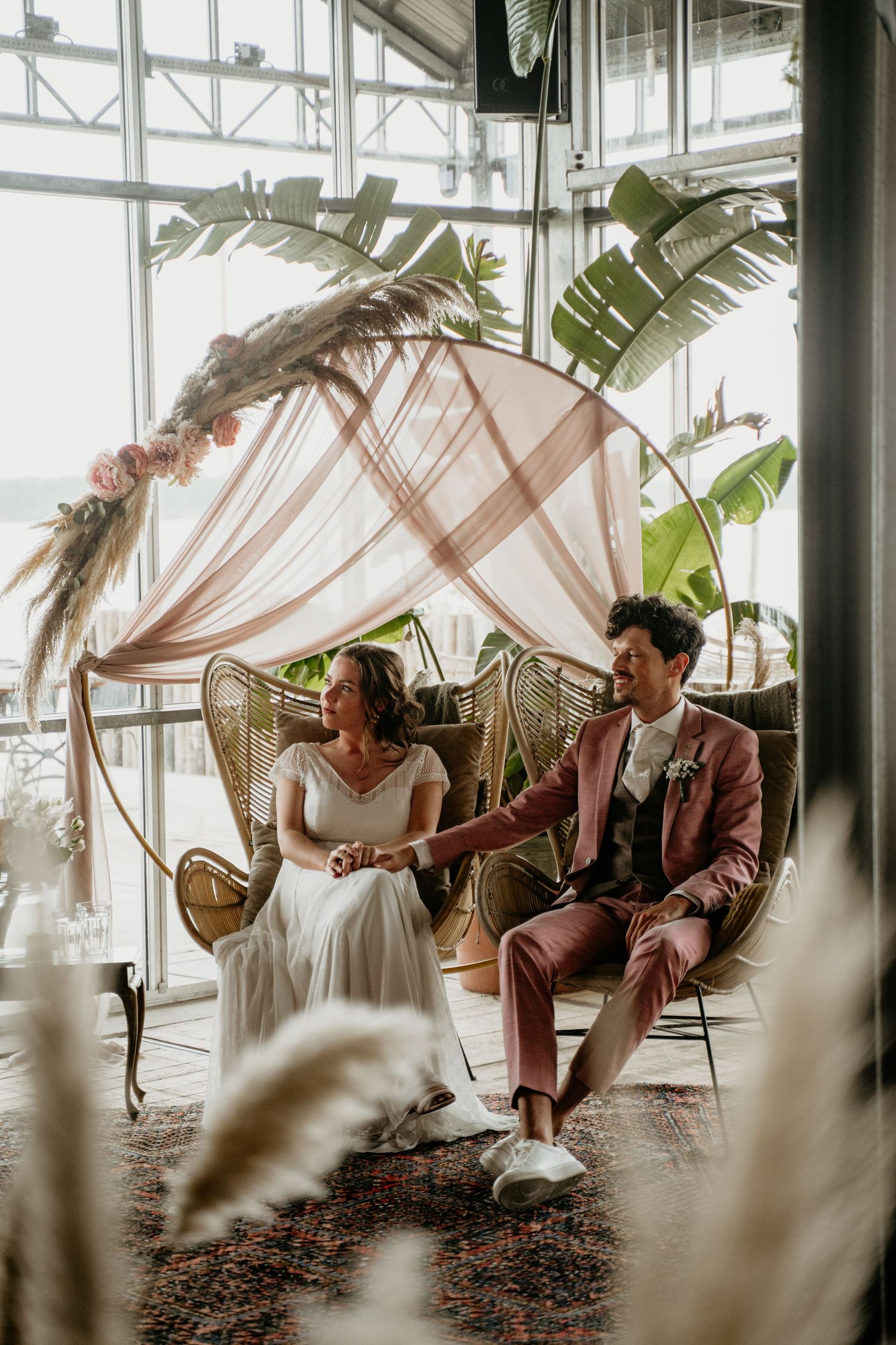 A romantic wedding setup with a blush canopy and vintage peacock chairs in a bright greenhouse setting.