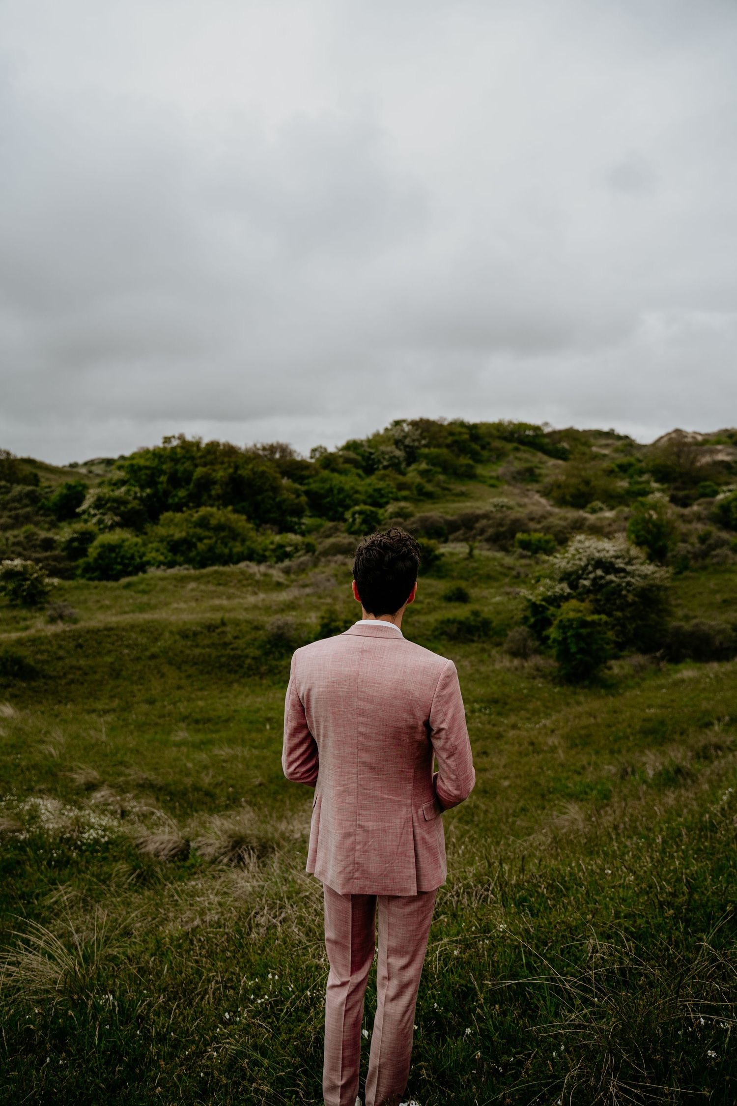 A person in a pink suit stands facing rolling green hills on an overcast day.