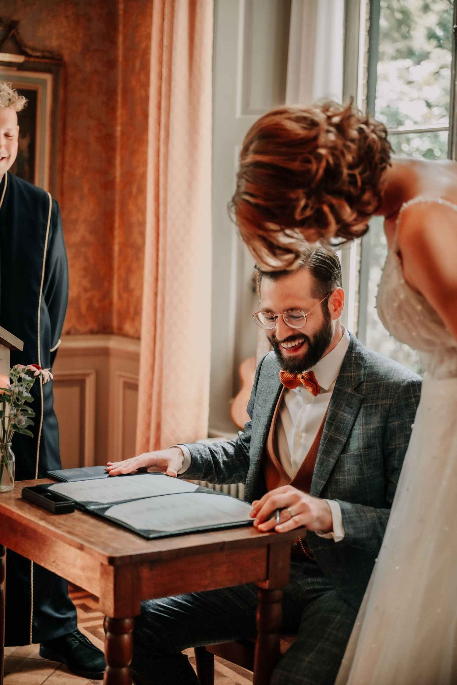 Man in grey suit and bow tie smiles while writing at wooden desk near window.