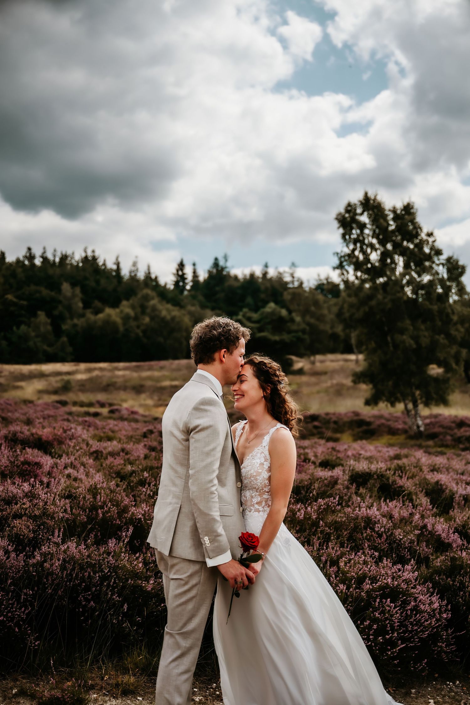A couple in wedding attire embraces in a field of purple heather under dramatic cloudy skies.