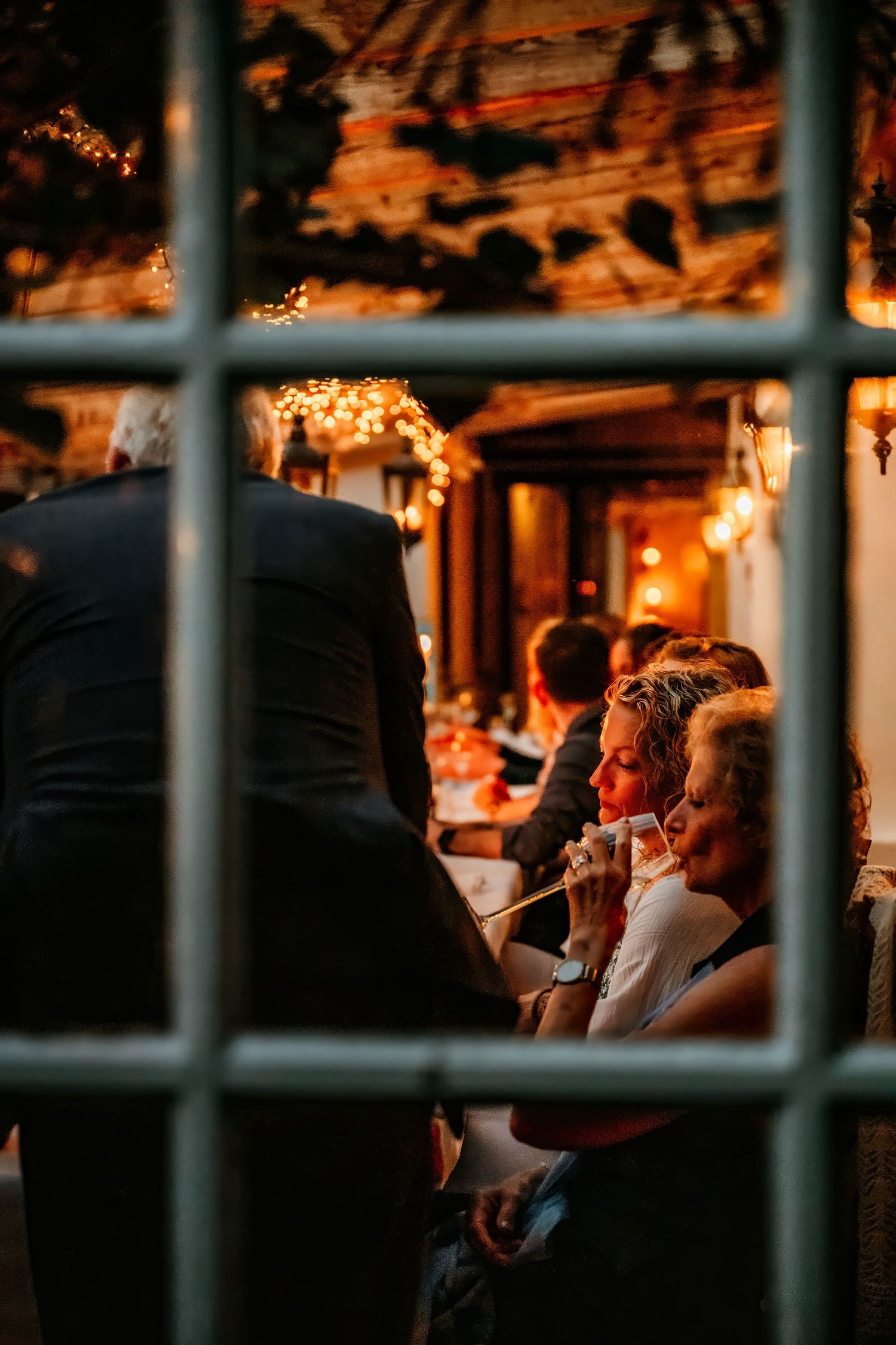 Warm candlelit reception venue viewed through window panes with guests enjoying evening celebrations.