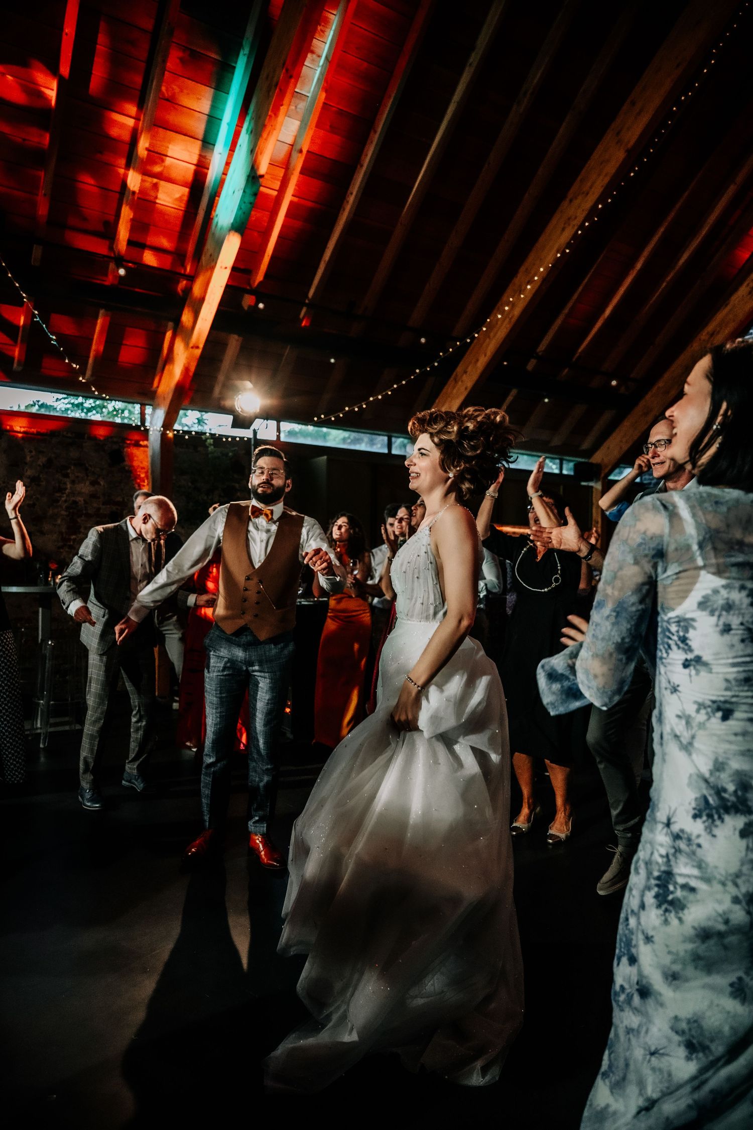 Wedding guests dance under colorful lights and string lighting at an evening reception.